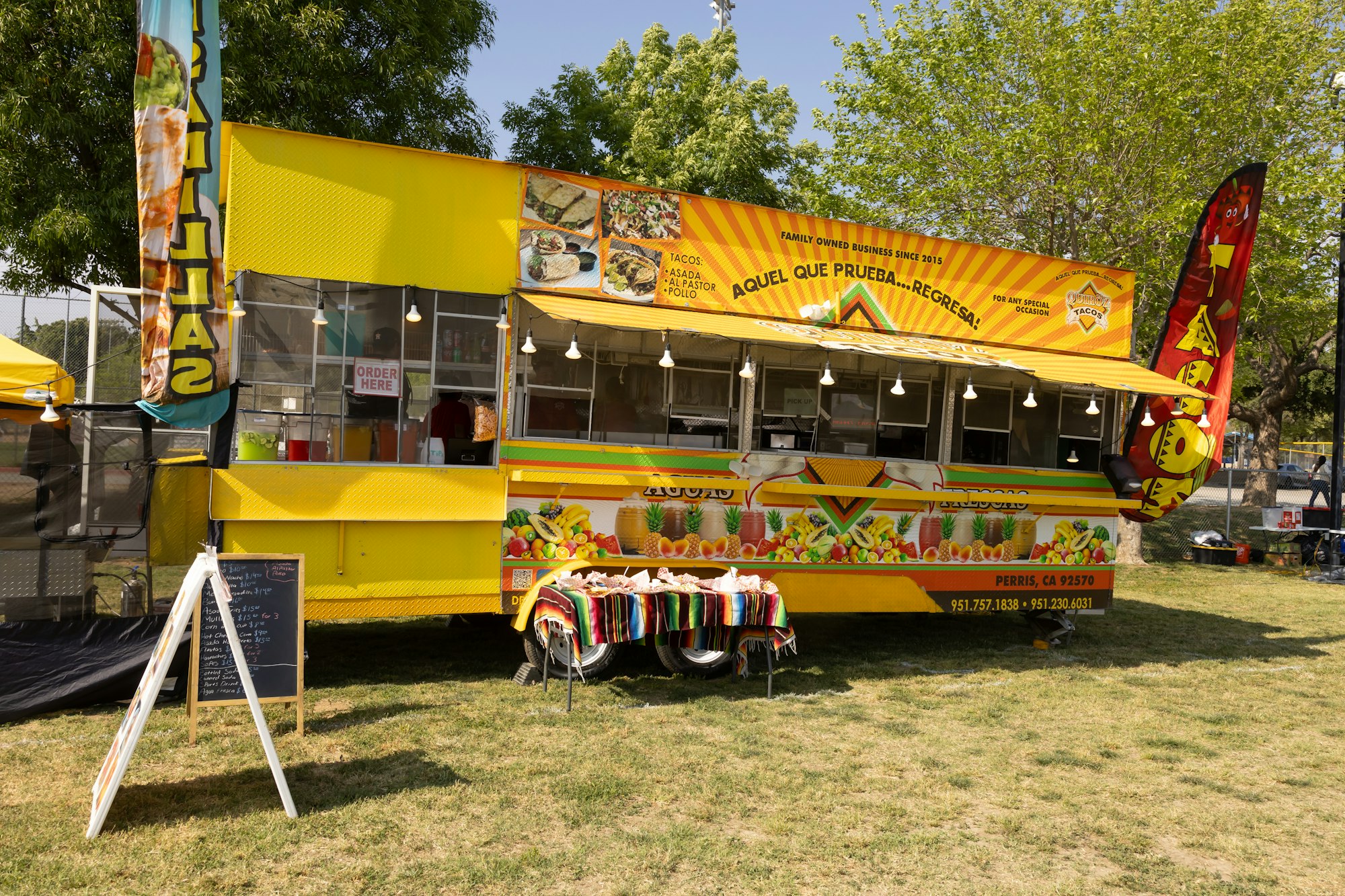 Yellow food truck with taco and fruit images, "Order Here" sign, open window, and outdoor setup with colorful tablecloth.