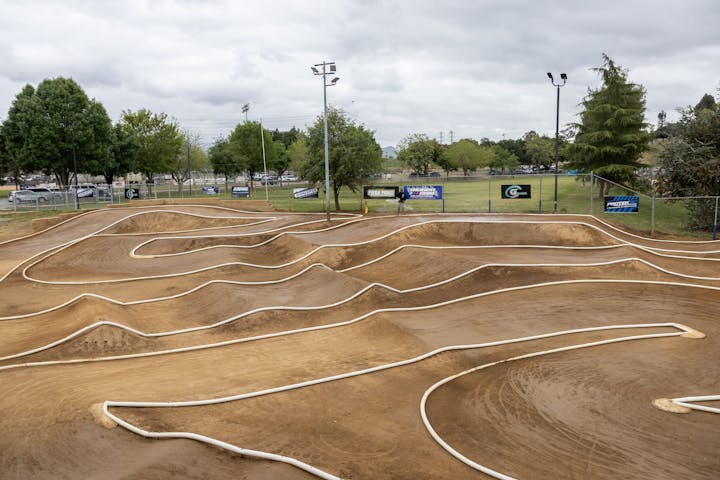 Outdoor RC car track with dirt paths and barriers, surrounded by trees and banners.