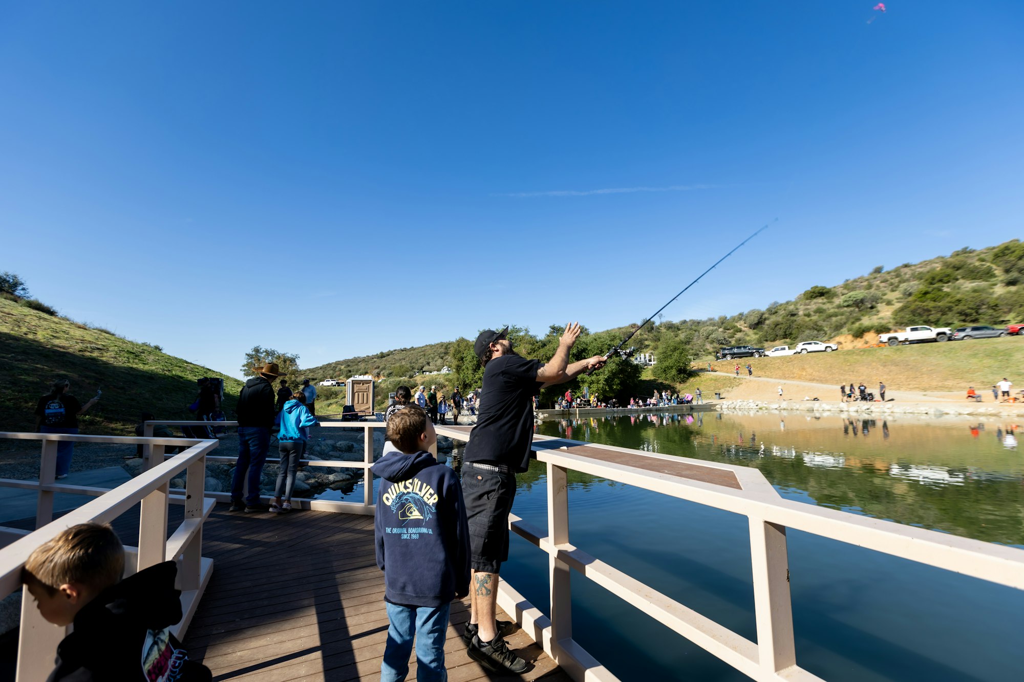 People fishing by a lake, surrounded by hills and cars parked on a dirt road.