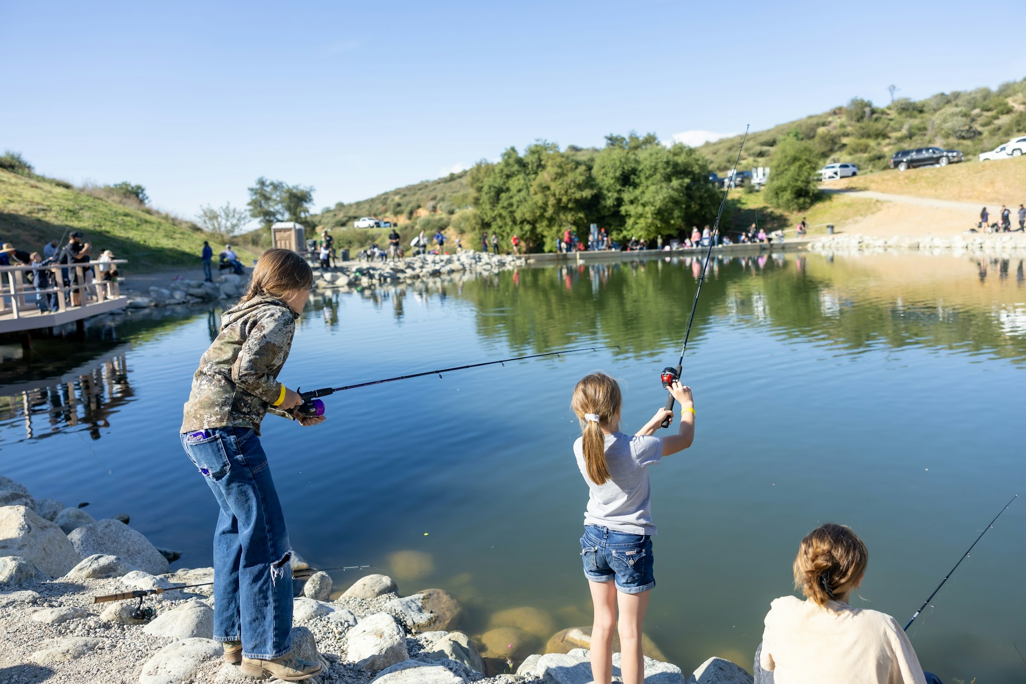 People fishing by a pond, with children holding fishing rods, others in the background, surrounded by trees and hills.