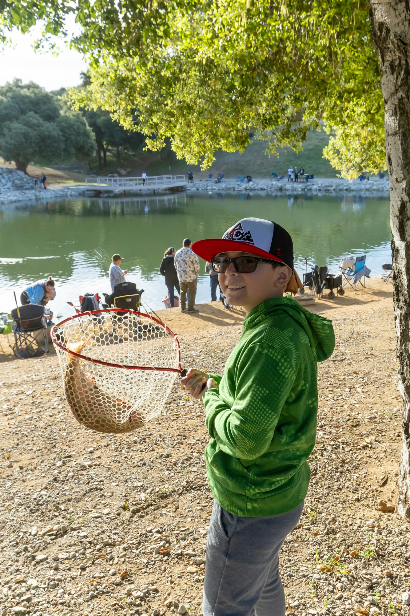 A child with a fishnet by a lake, holding a caught fish, surrounded by people fishing near the water.