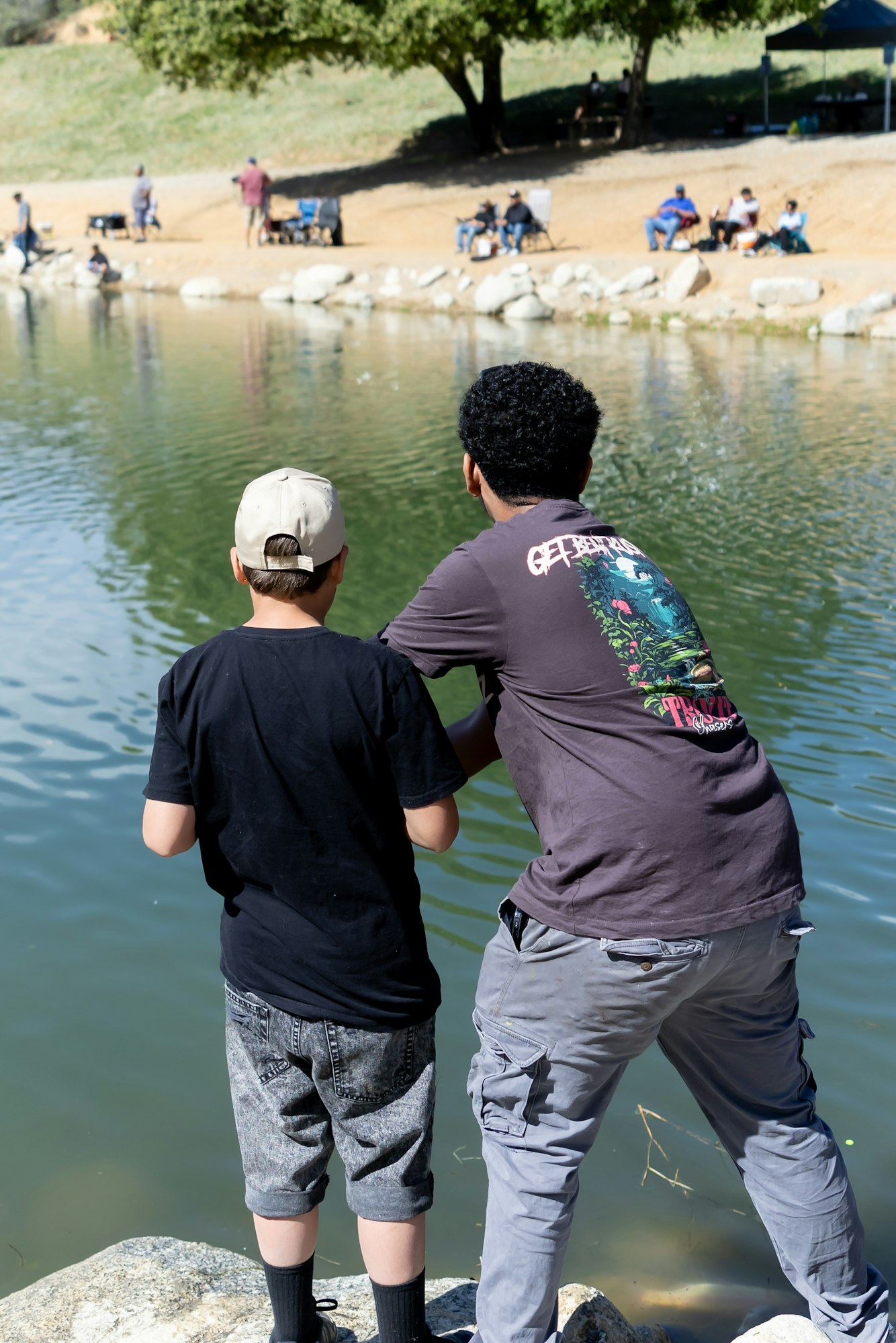 Two people fishing by a pond with others sitting and relaxing in the background.