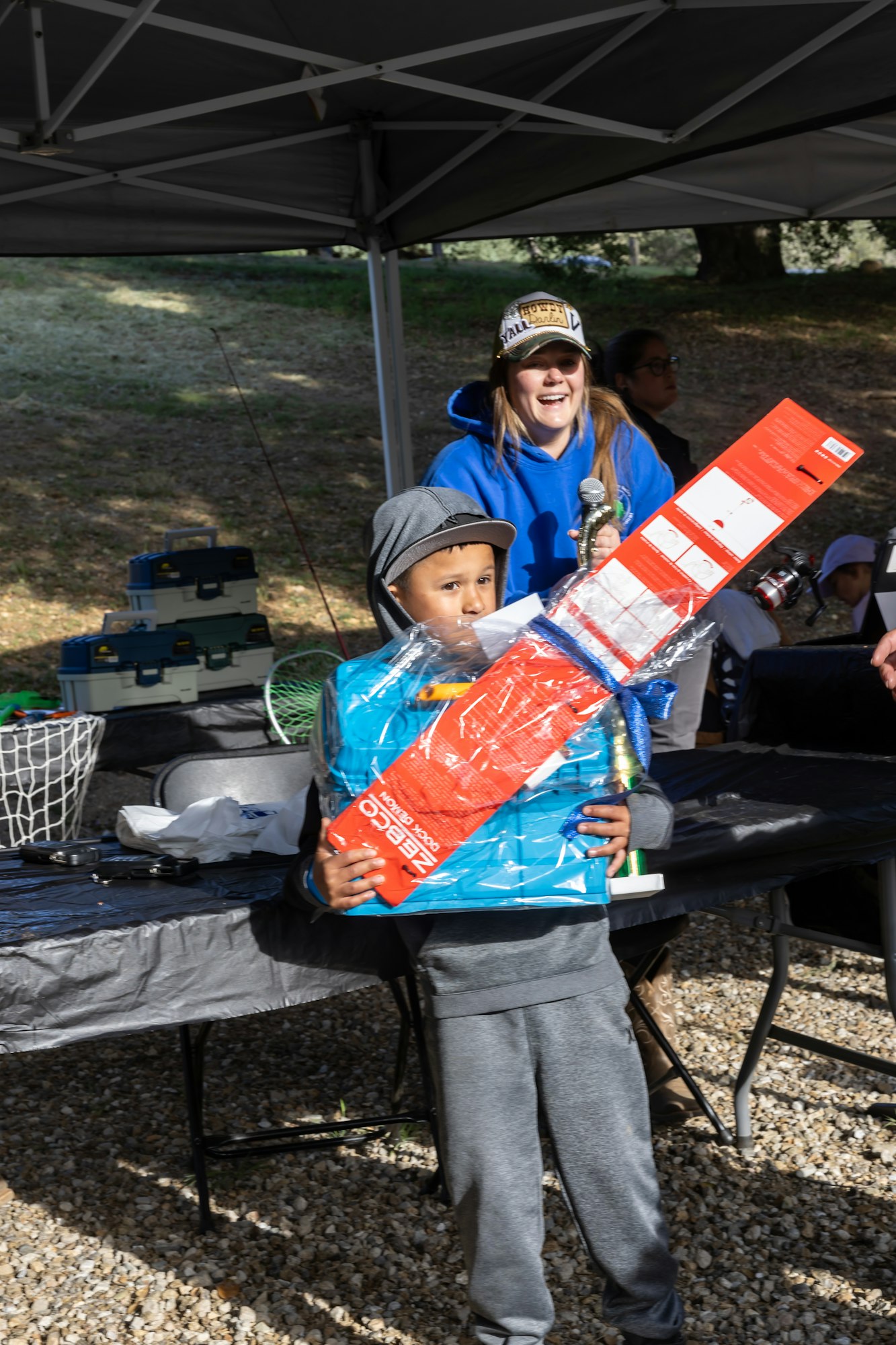 A child holds a Zebco fishing rod and tackle box under a canopy, with a person behind holding a microphone.