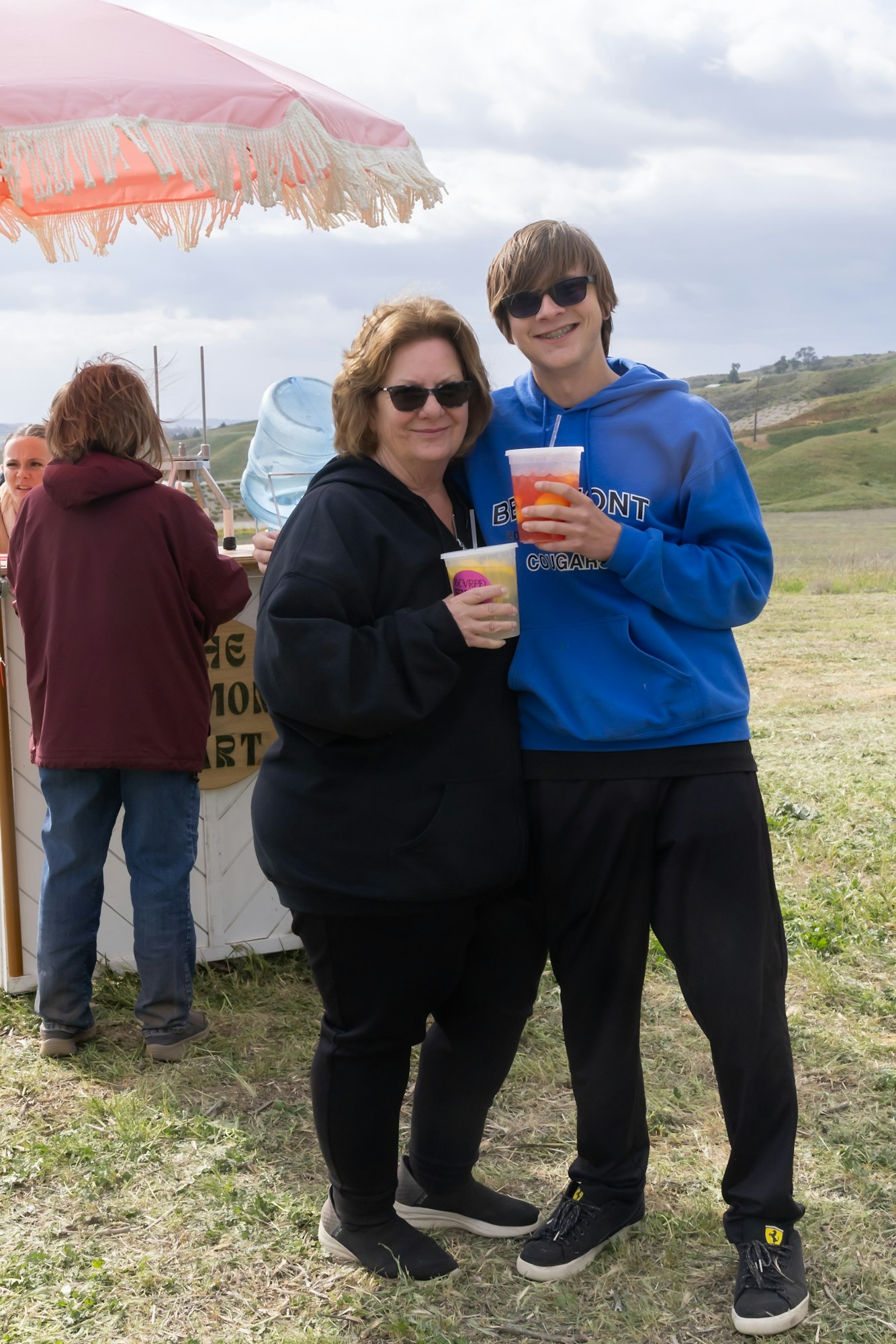 Two people with drinks, smiling outdoors near a vendor booth and umbrella.