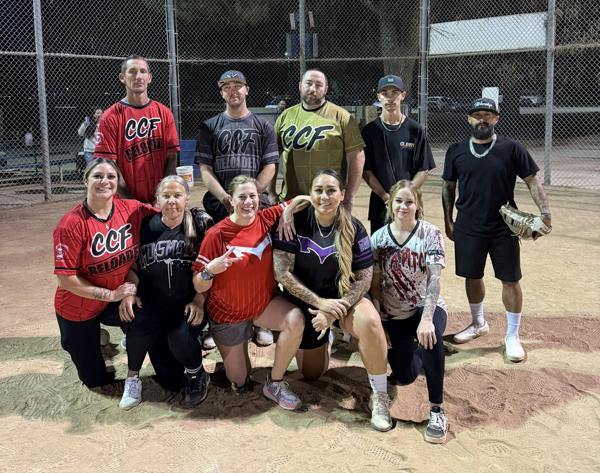 A group of people in sports uniforms posing on a baseball field, some kneeling, others standing.