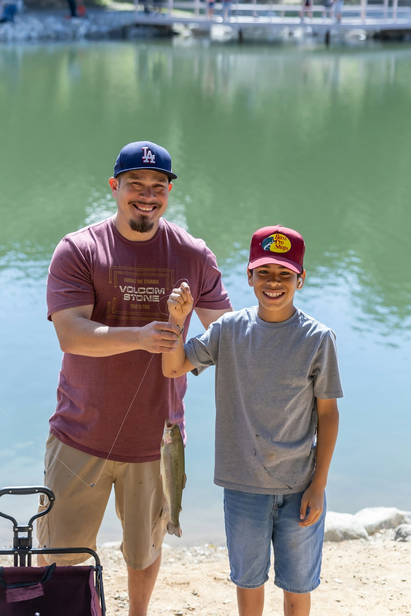 Two people smiling by a lake, holding a fish they caught.