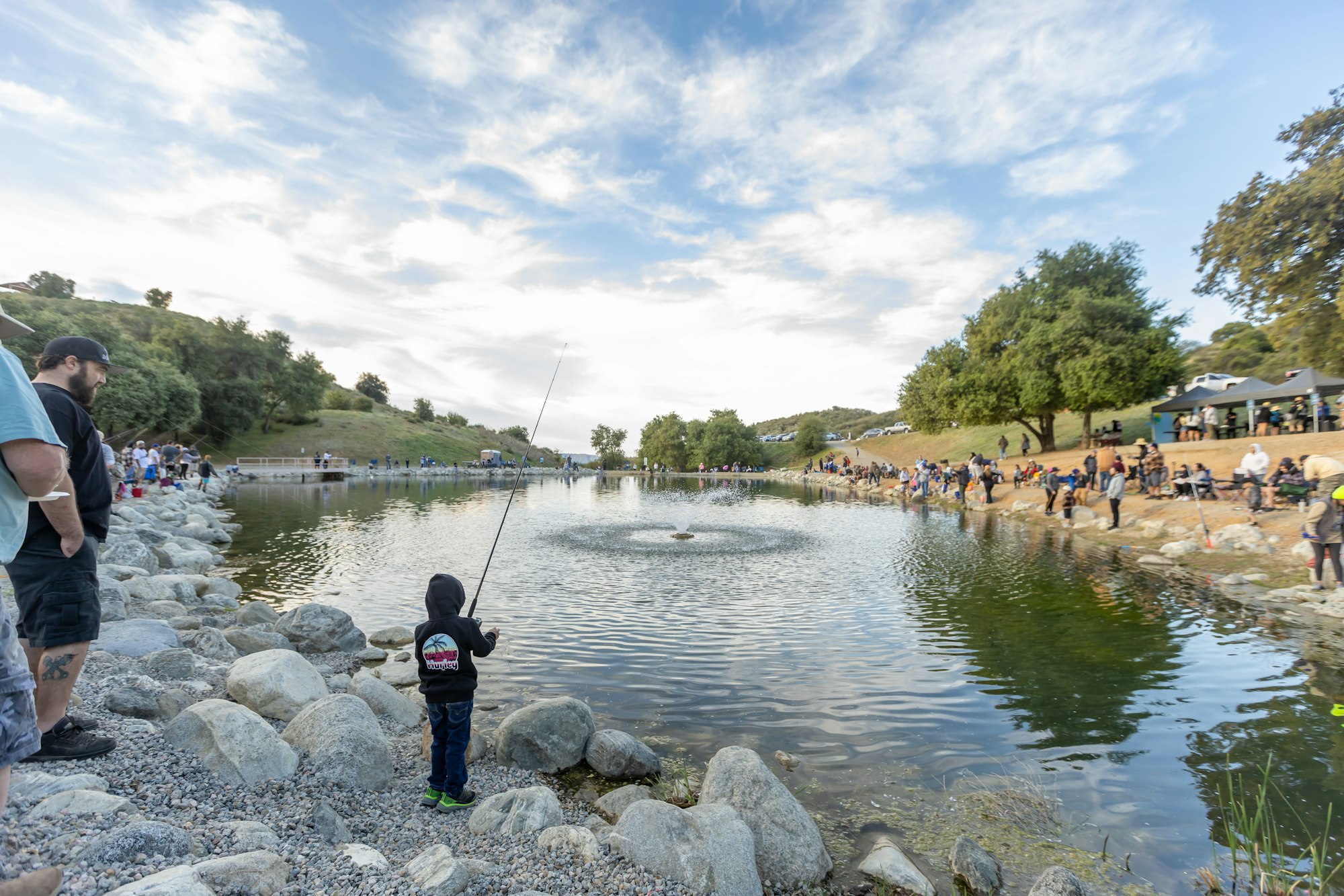 People fishing at a lake with a scenic, cloudy sky, surrounded by trees and rocks.