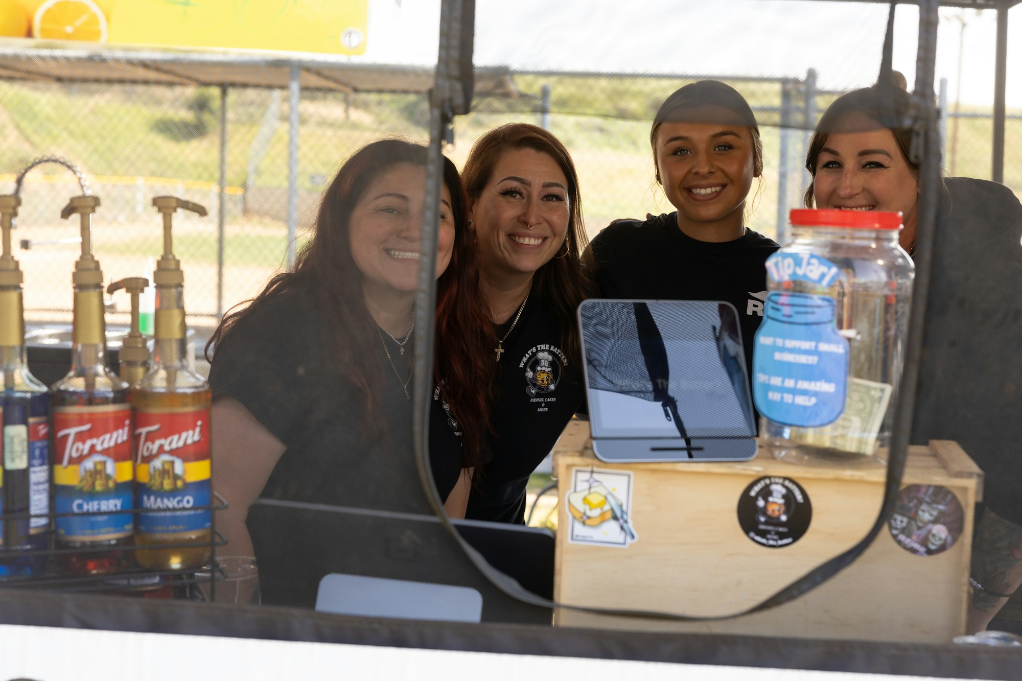 Four people smiling at a booth with syrup bottles and tip jar, seen through a window.