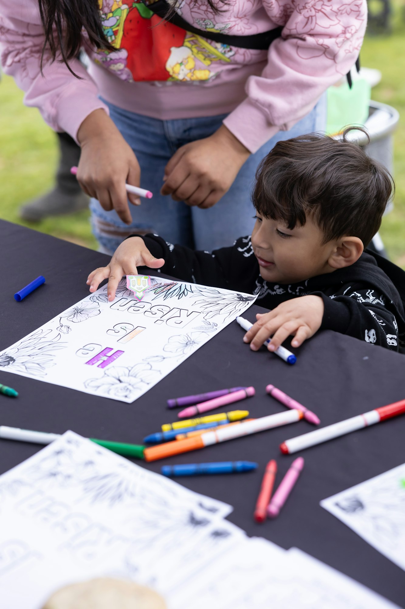 A child is coloring a page with crayons and markers on a table, supervised by an adult.