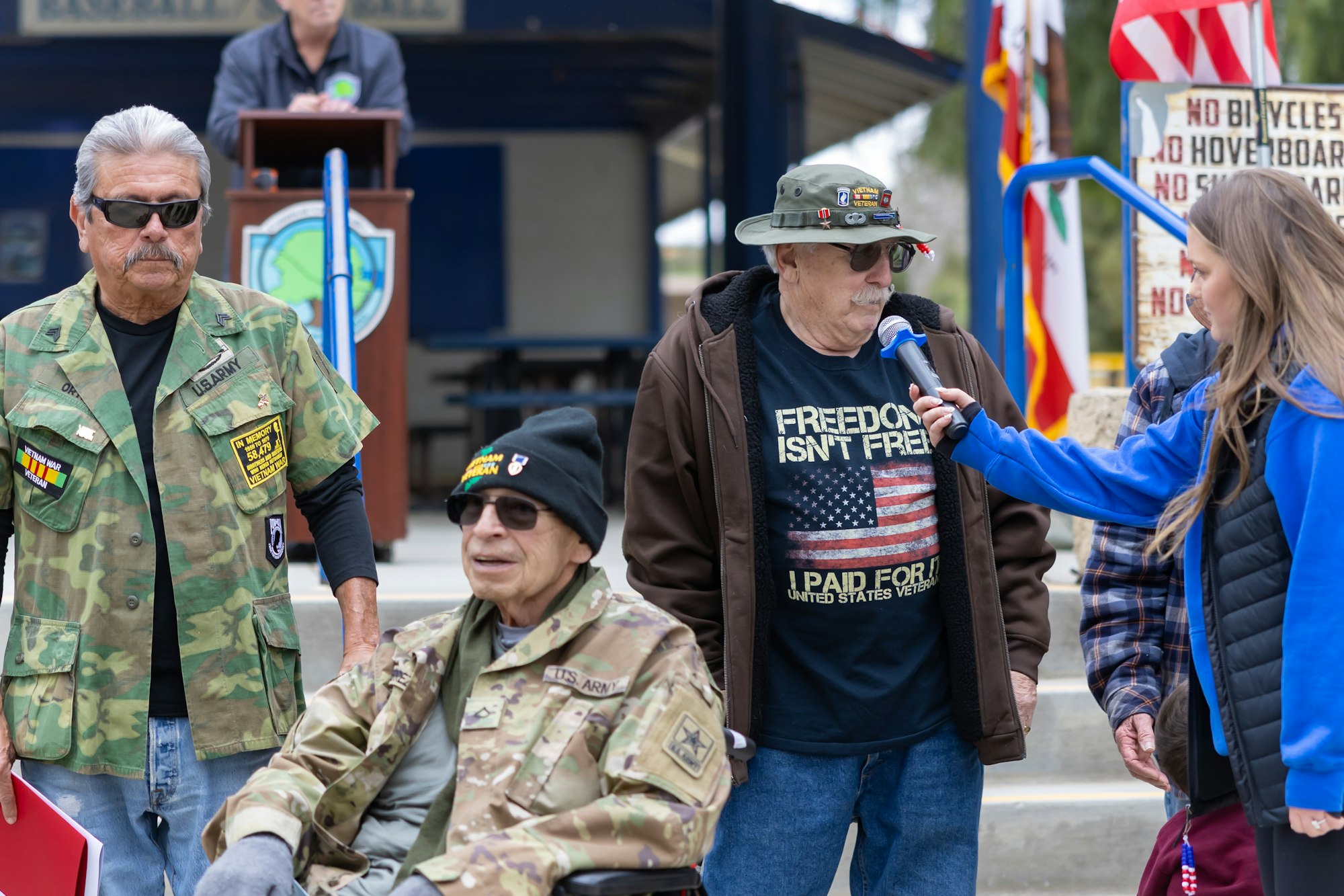 A veteran event with people in military attire, one speaking into a microphone held by a woman. Signs and flags are visible in the background.