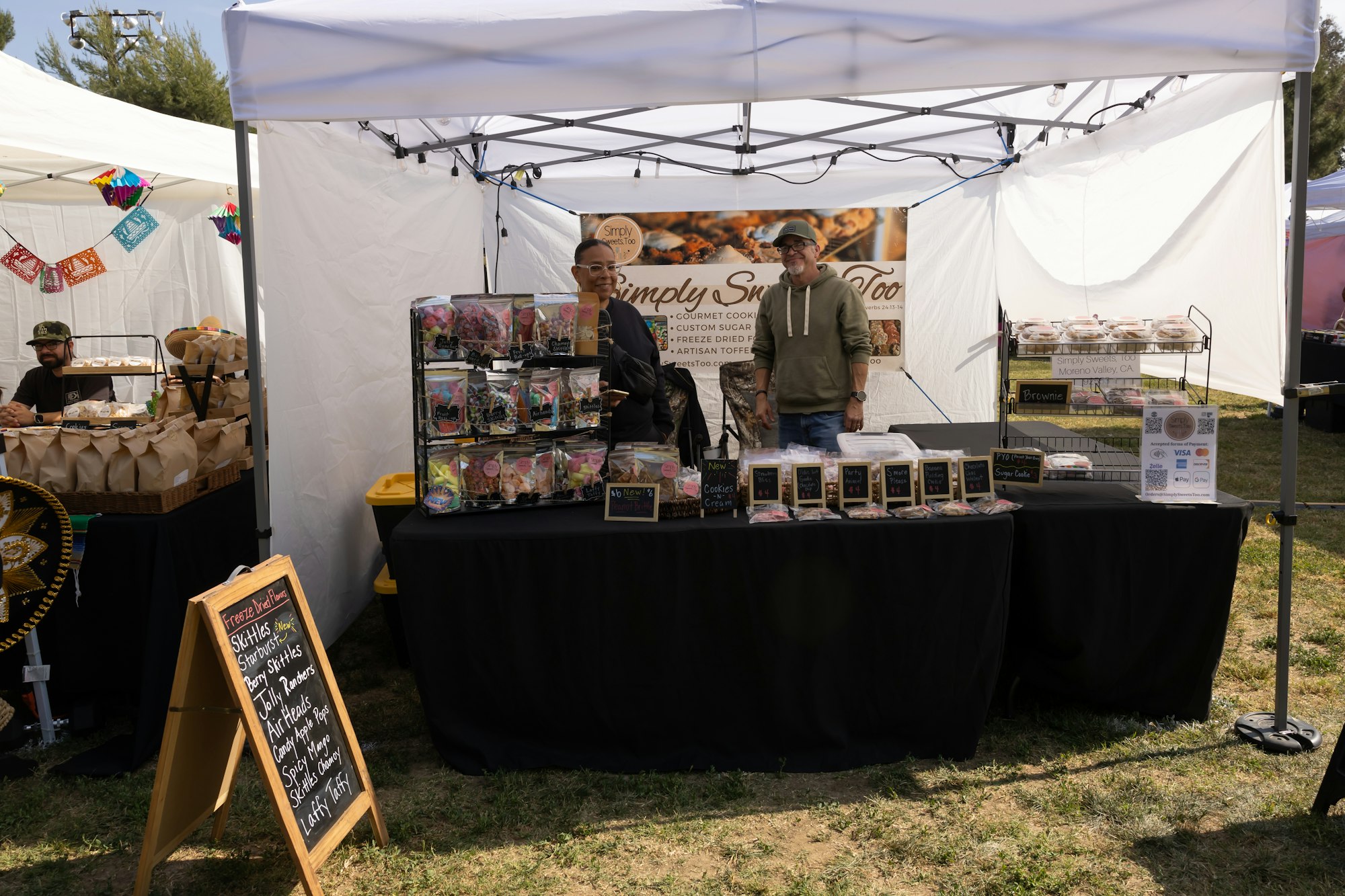 Market stall selling gourmet cookies and treats, with two people standing behind a display and a sign listing various snack options.