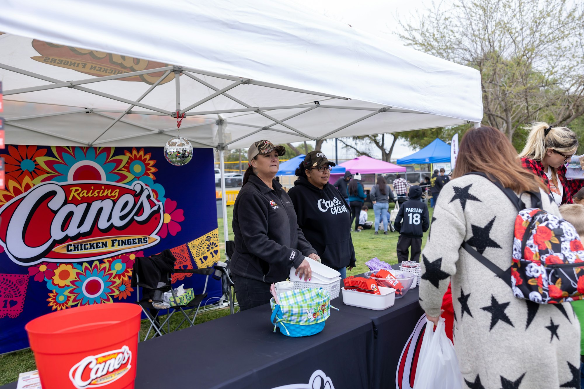 People at a Raising Cane's Chicken Fingers booth at an outdoor event, under a tent with bright decorations.