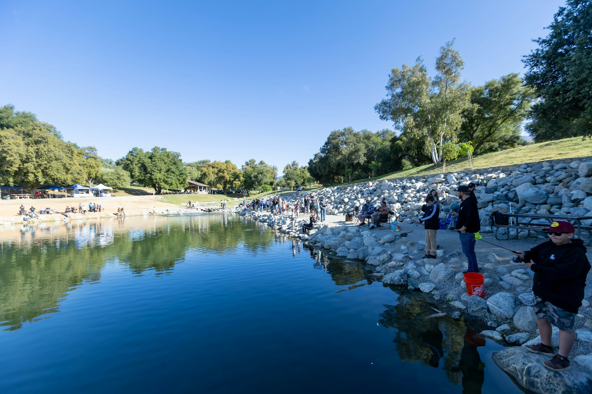 People fishing by a rocky lakeside under a clear blue sky, surrounded by trees and picnic tables.