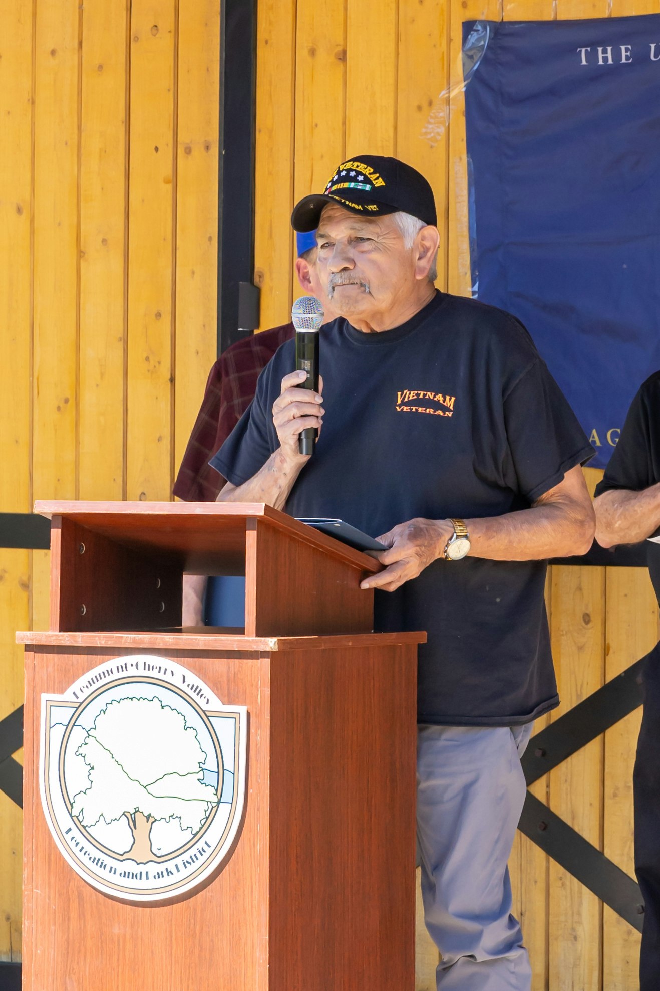 A man speaking at a podium, wearing a Vietnam veteran cap, during an event with a wooden backdrop.