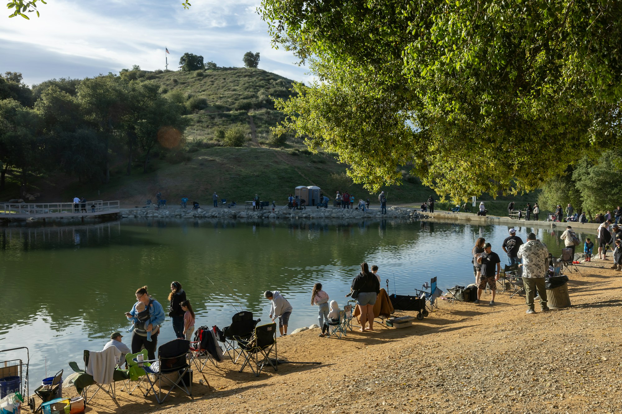 People fishing and relaxing by a lakeside, with chairs and fishing gear, under trees and a hill with a flag in the background.