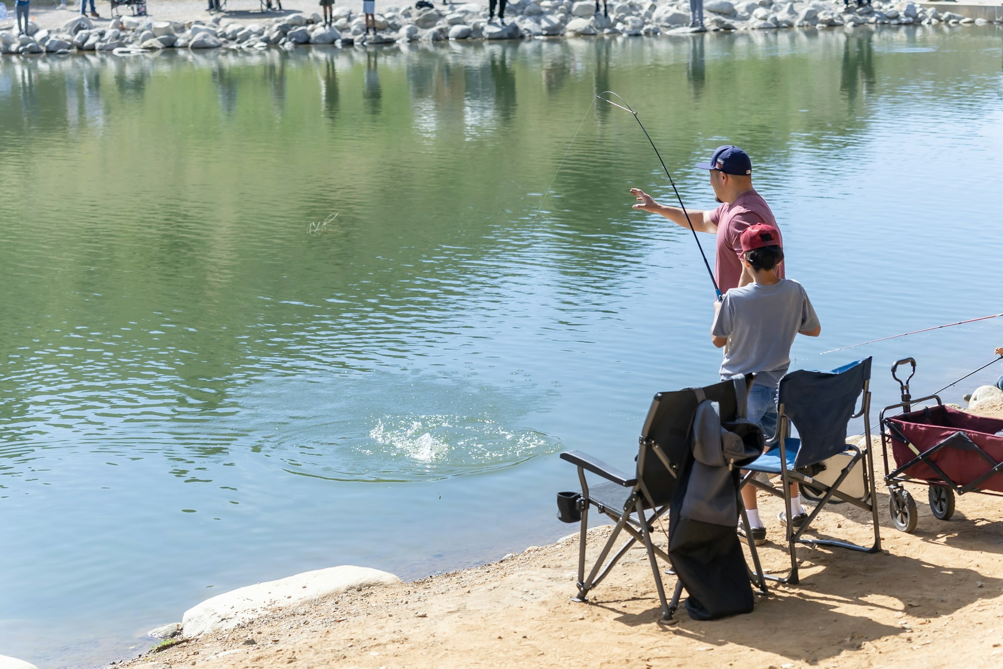 Two people are fishing by a lake with chairs and a wagon nearby.