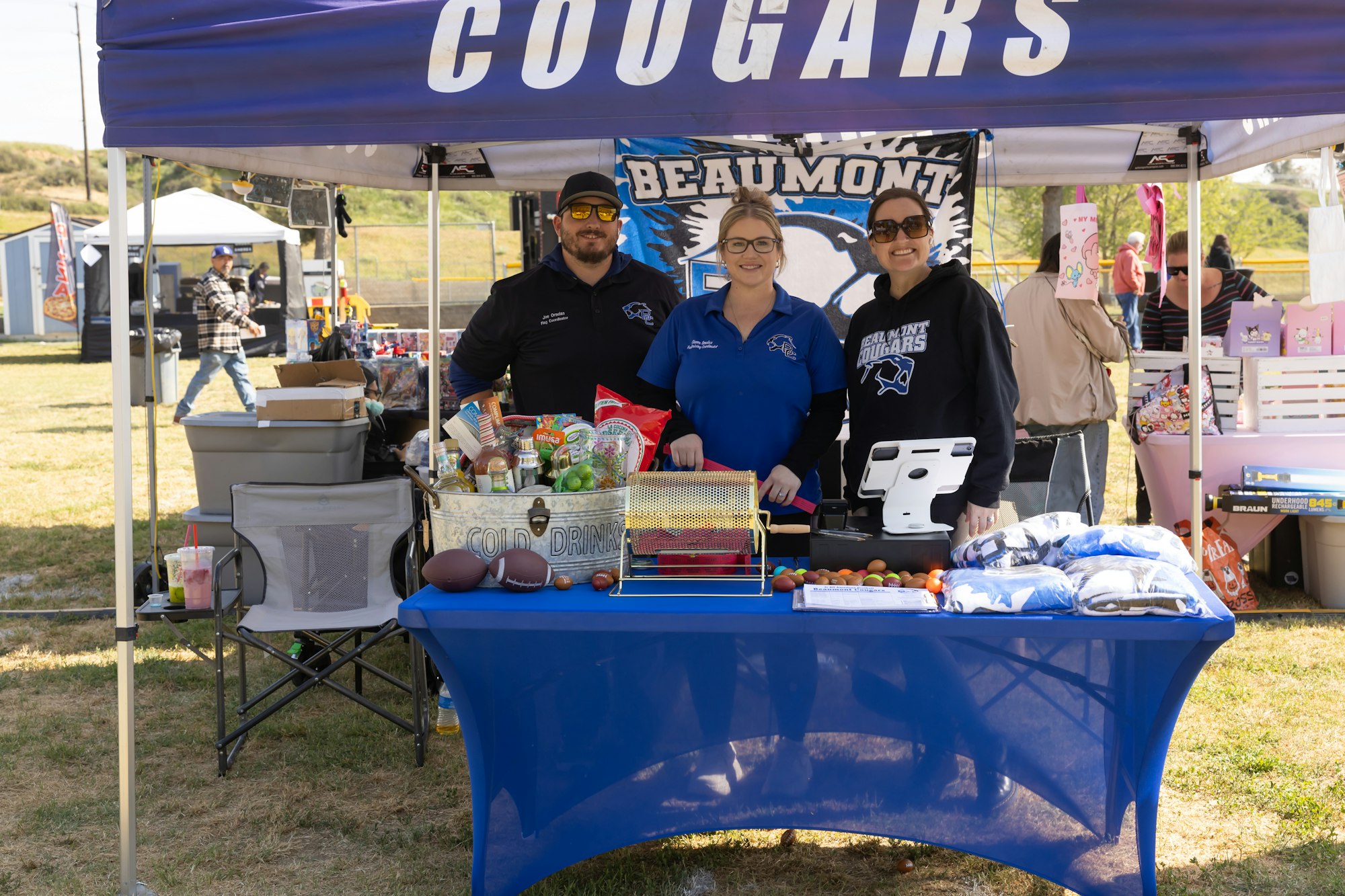 Three people stand under a "Cougars" tent, selling items on a blue table at an outdoor event.