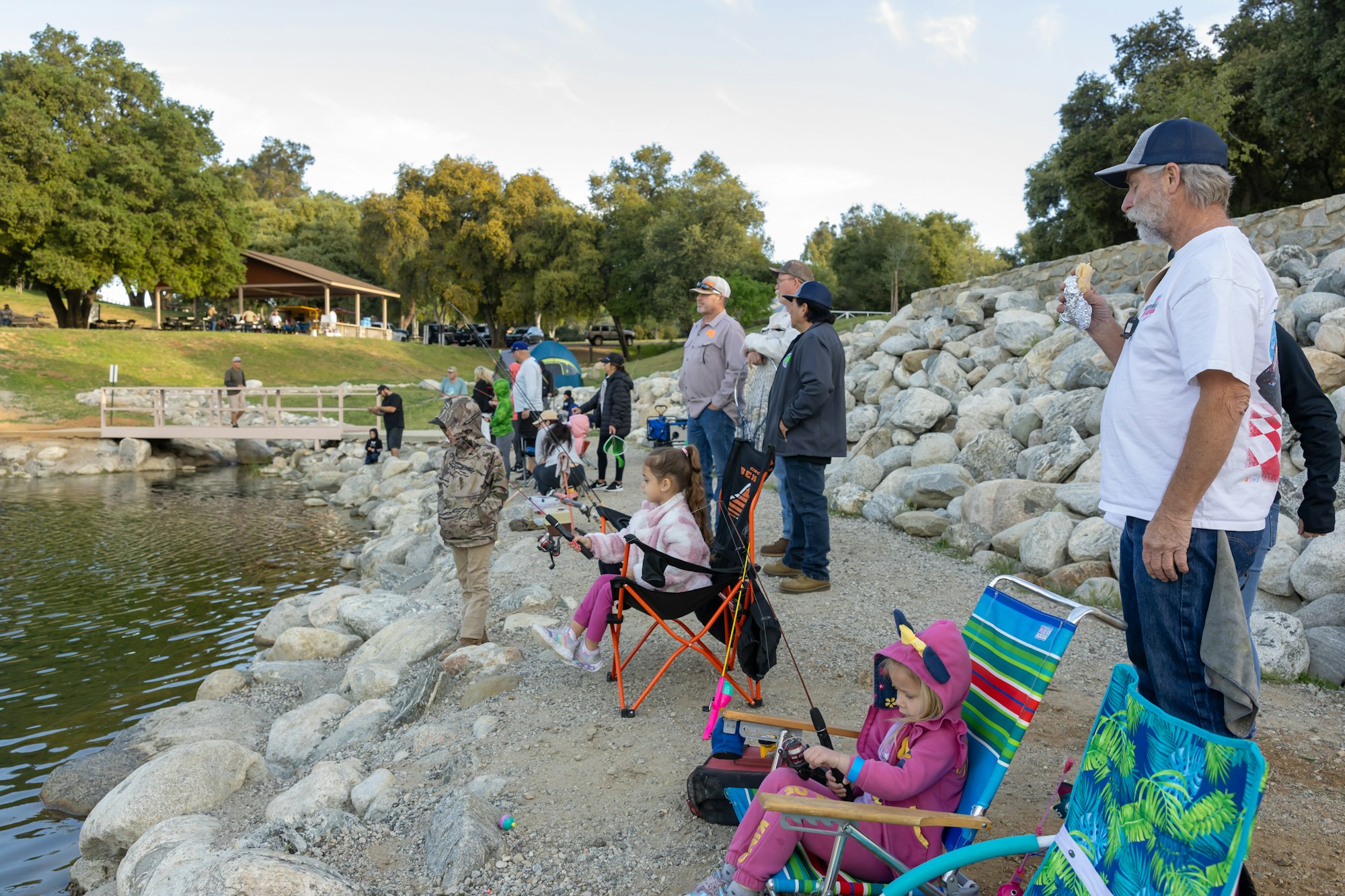 People fishing by a lake; adults and children sitting or standing with rods.