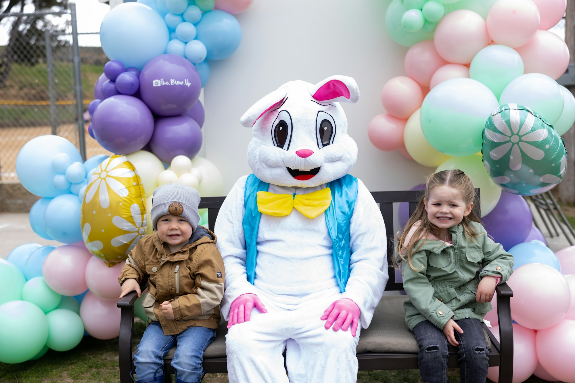 Two kids sit with a person in an Easter bunny costume, surrounded by colorful balloons.