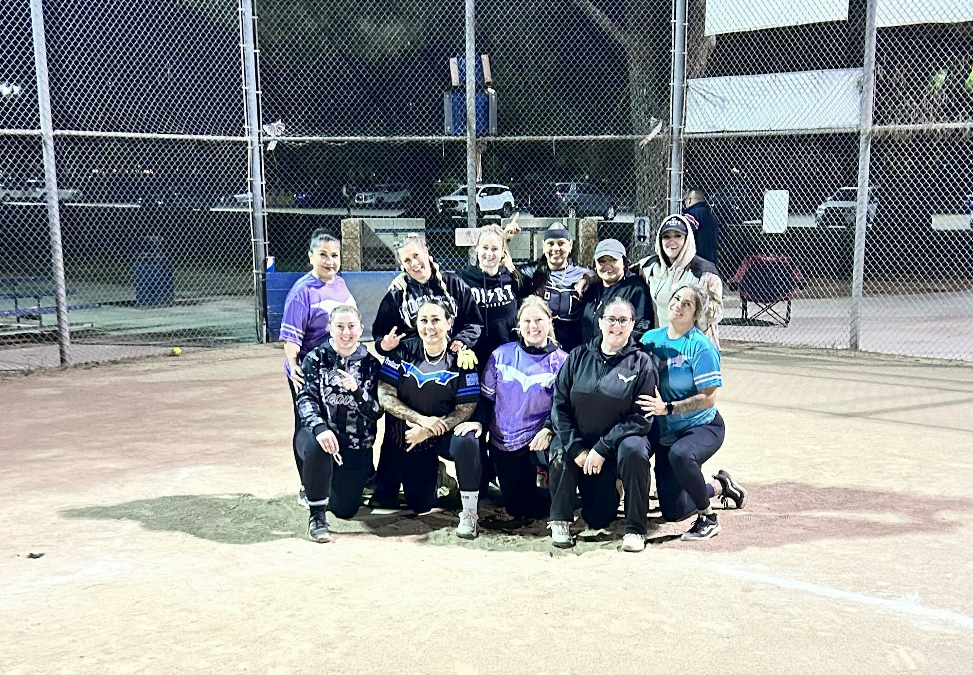 A group of people posing on a softball field at night, with a fence and vehicles in the background.