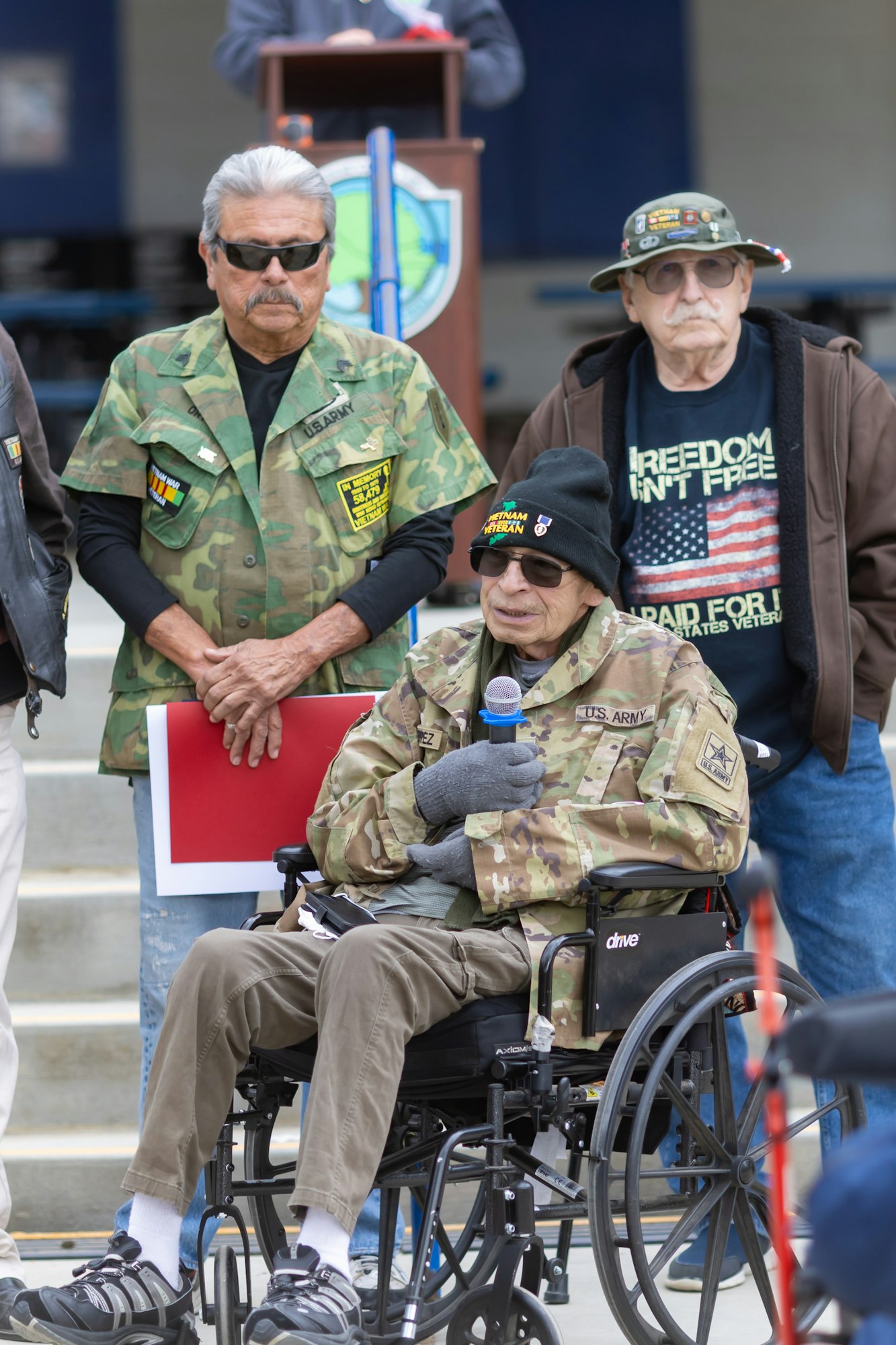 Veterans in military attire, with one in a wheelchair speaking into a microphone.