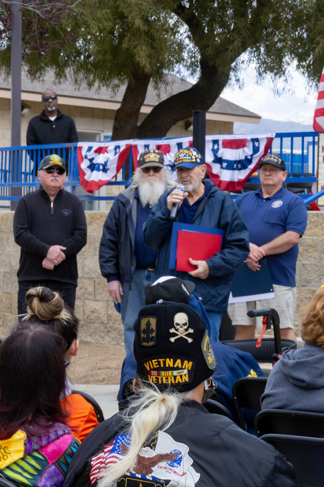 A group of people, some wearing military caps, during a speech at an outdoor event with patriotic decor.