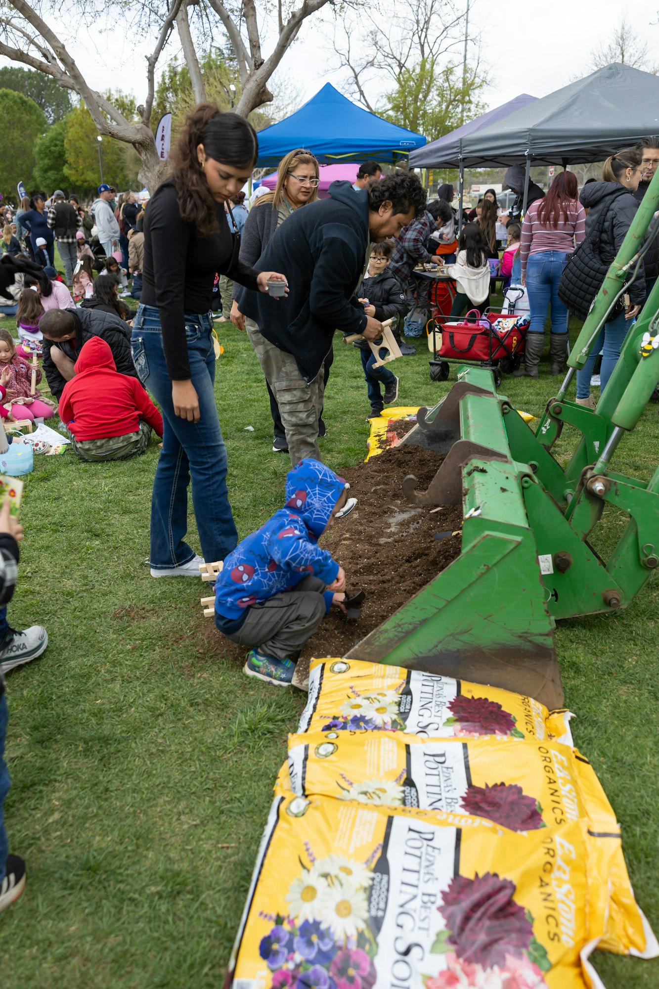 A crowd at an outdoor event, with people digging in soil near potting soil bags and tents in the background.