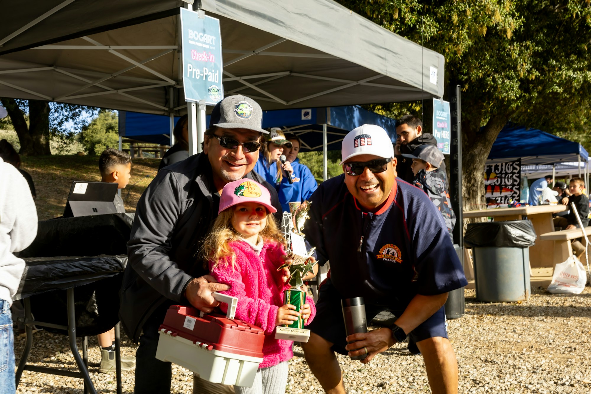 A child holding a trophy with two adults, under a canopy labeled “Check-In Pre-Paid”, at an outdoor event.