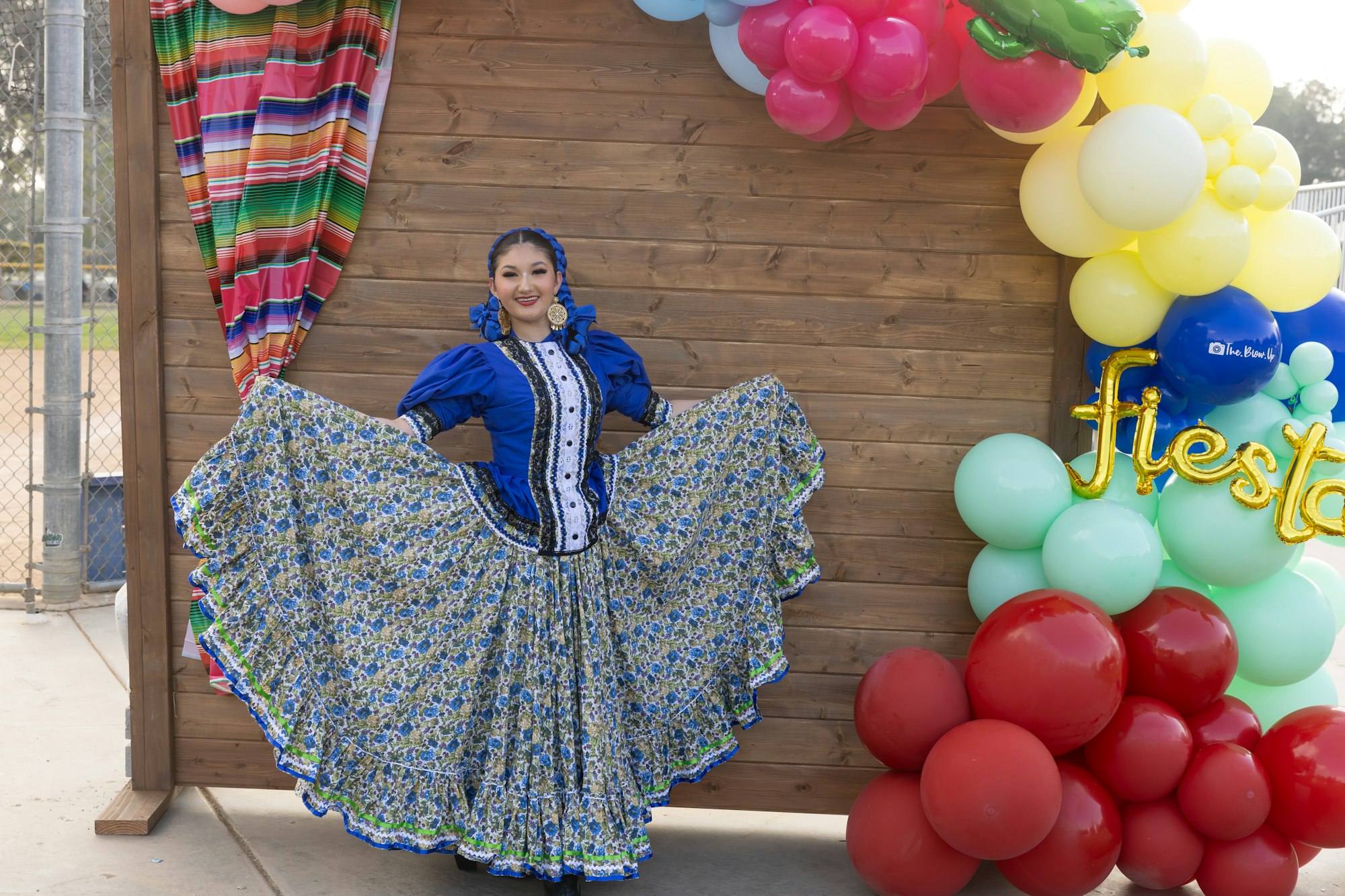 Person in traditional blue dress posing at a fiesta-themed photo spot with colorful balloons and decorations.