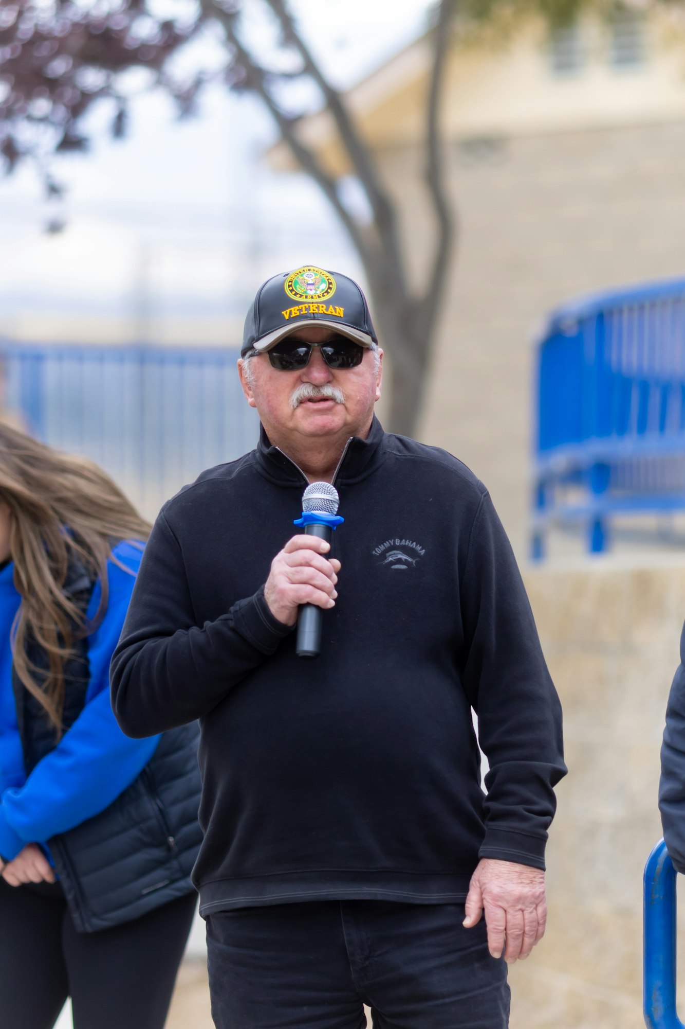 A person wearing a "Veteran" cap and sunglasses is speaking into a microphone outdoors.