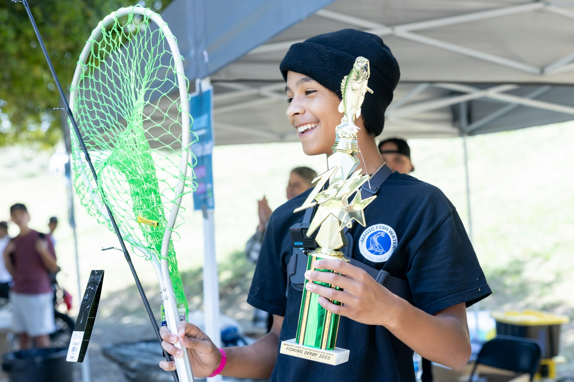 A person smiles, holding a fishing trophy and a net under a tent.