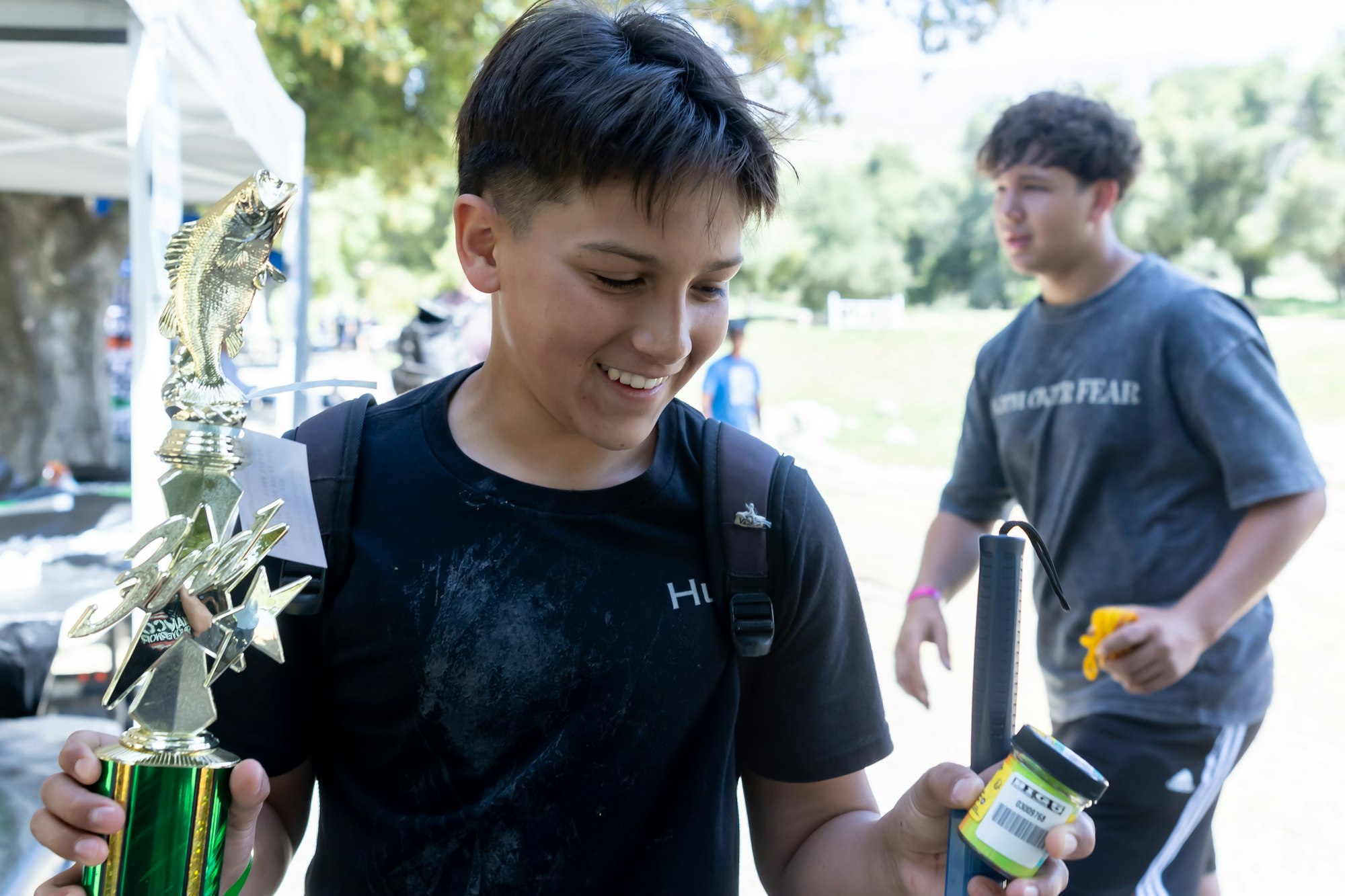 A person smiles holding a fish trophy and a small container, with another person blurred in the background outdoors.