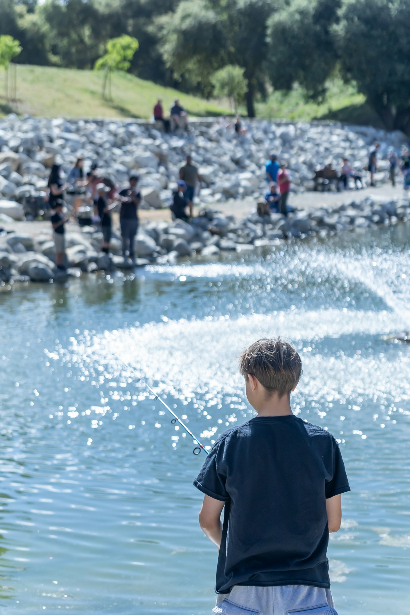 A person fishing at a pond with a fountain, surrounded by people on a rocky shore.
