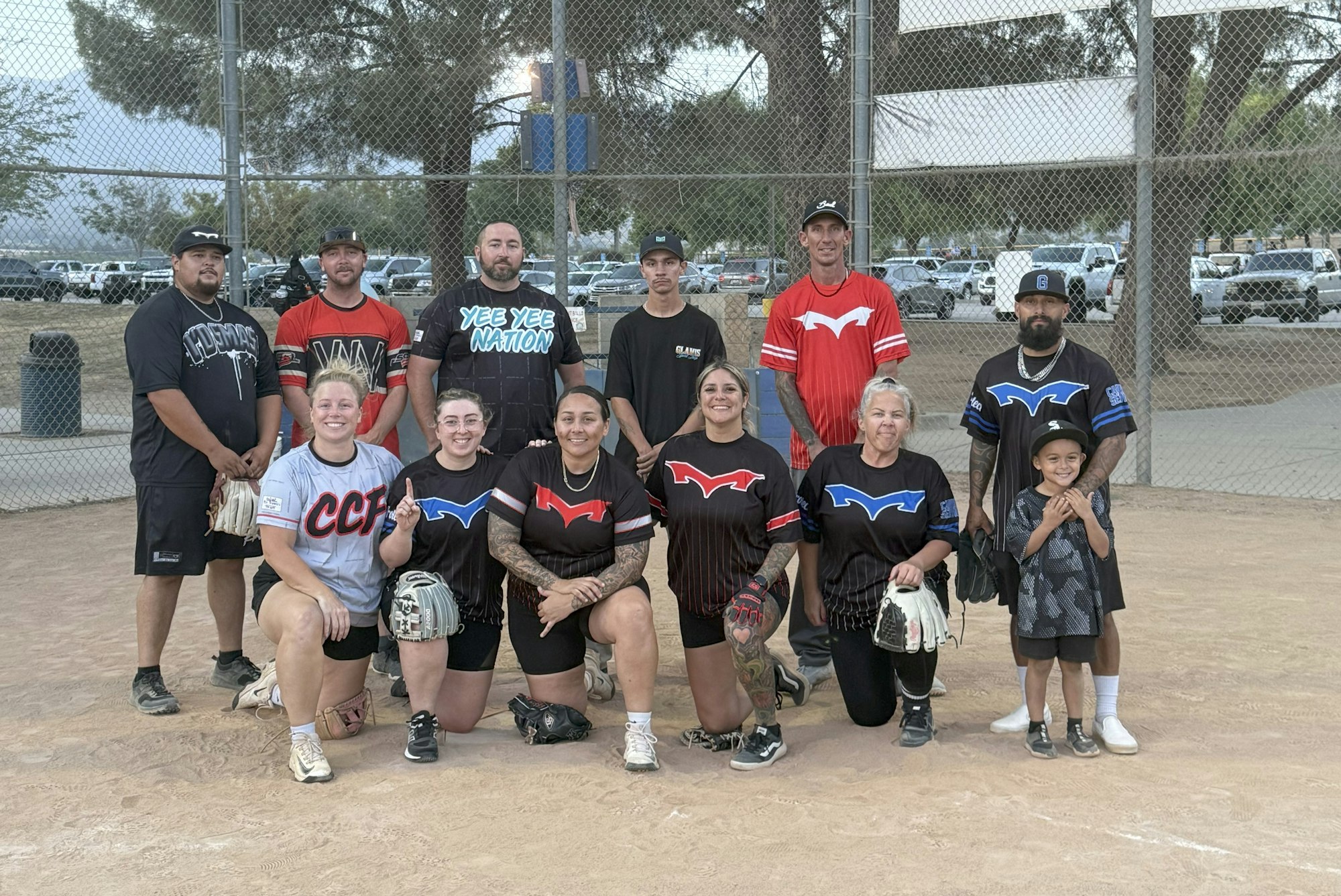A group of people posing on a baseball field, some kneeling, wearing different team shirts, with a chain-link fence in the background.