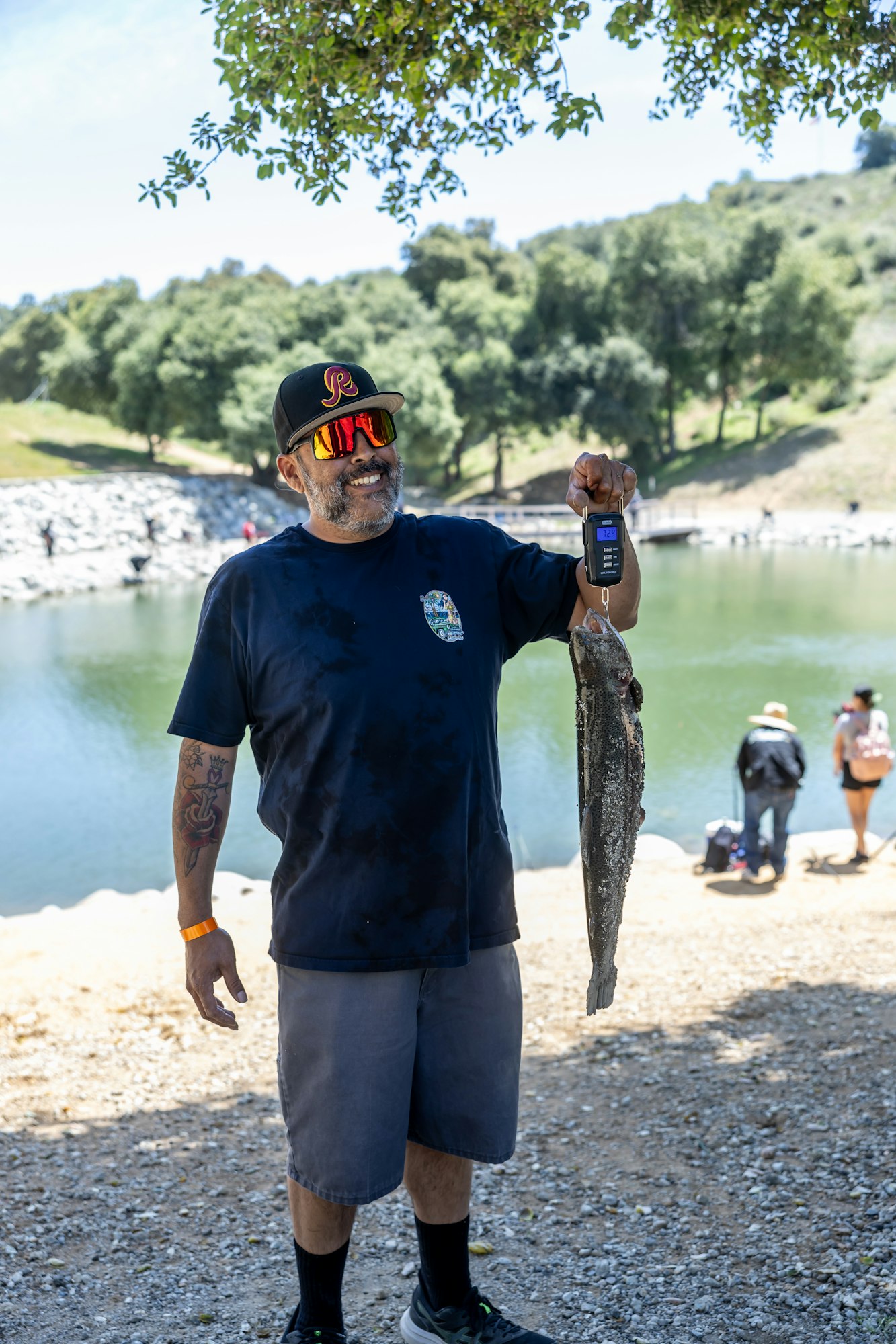 A person holding a large fish on a digital scale, standing by a lake, wearing sunglasses and a cap.