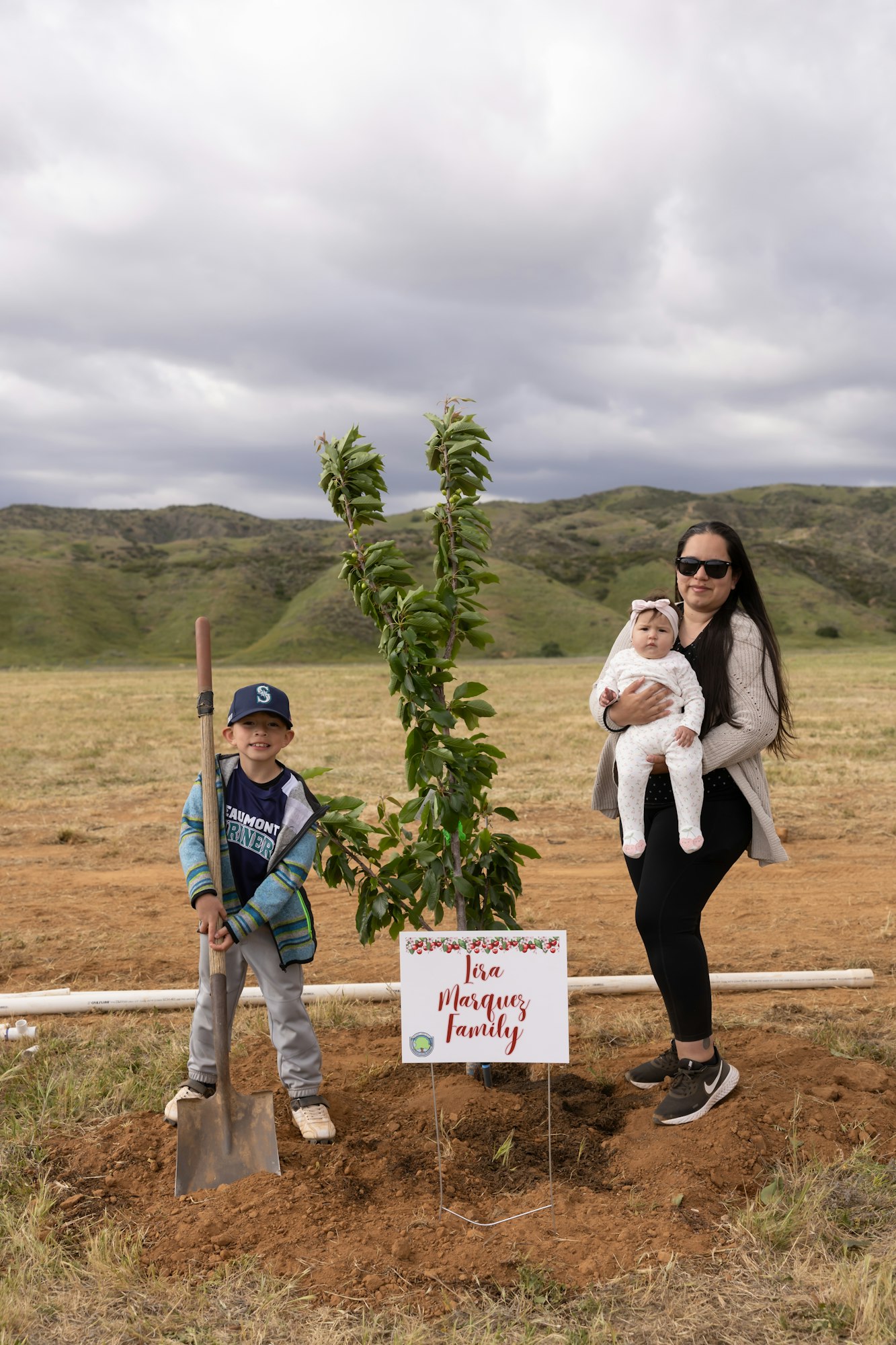 A woman with a baby and a child holding a shovel stand next to a planted tree with a sign reading "In Marquez Family."