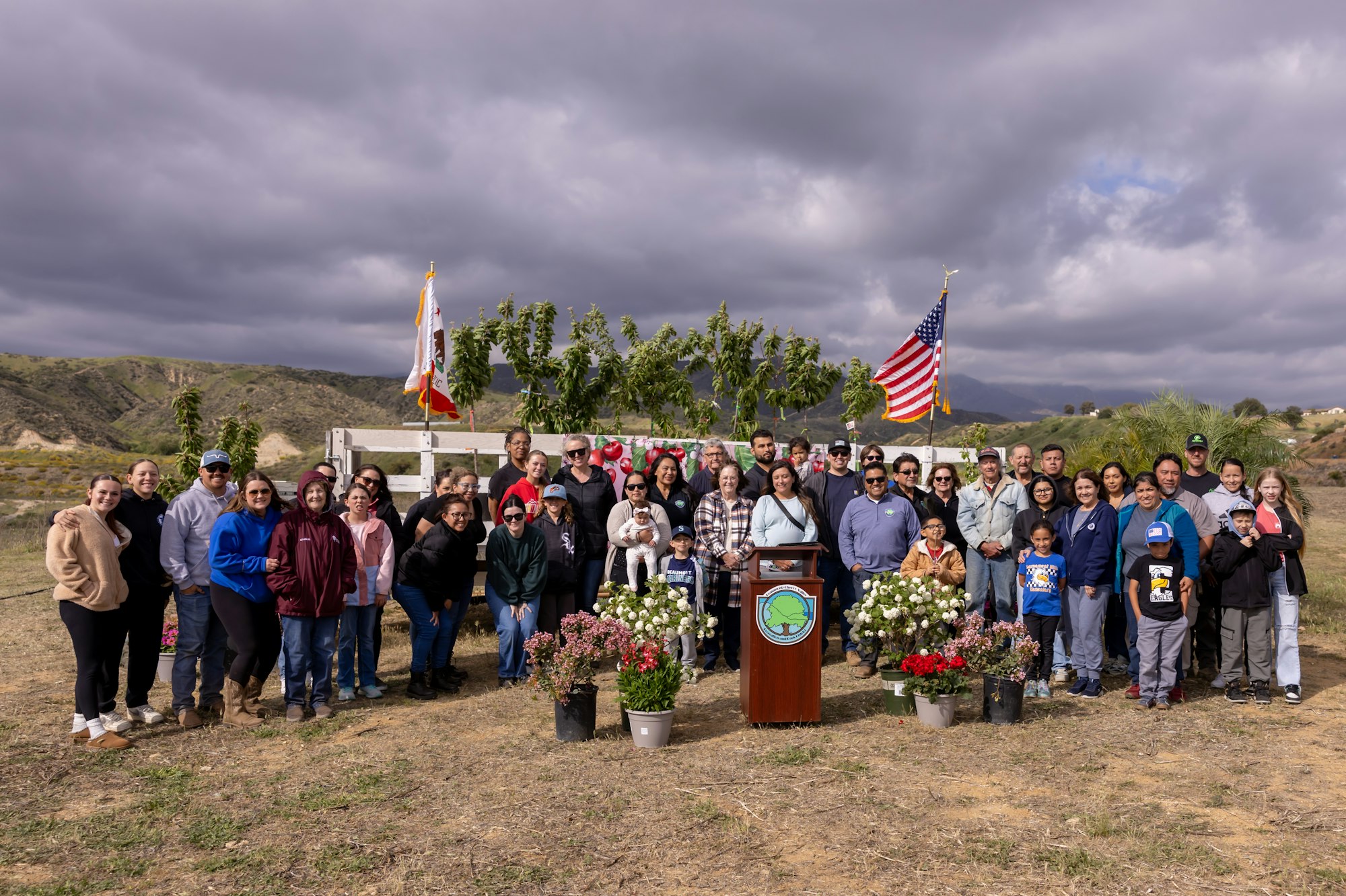 A group of people gathered outdoors near flags and plants under a cloudy sky, with hills in the background.