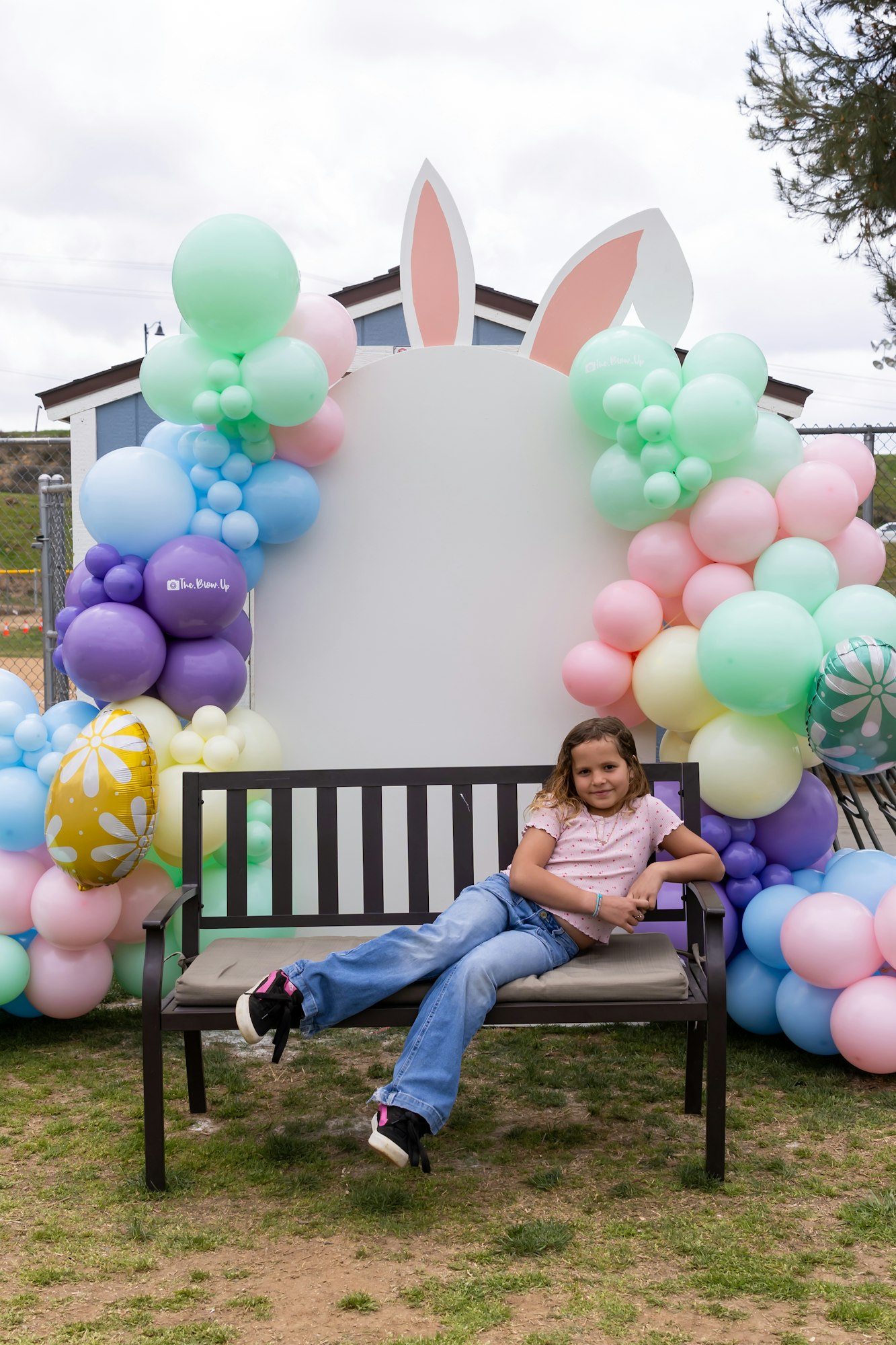 A child sitting on a bench surrounded by colorful balloons with bunny ears in the background.
