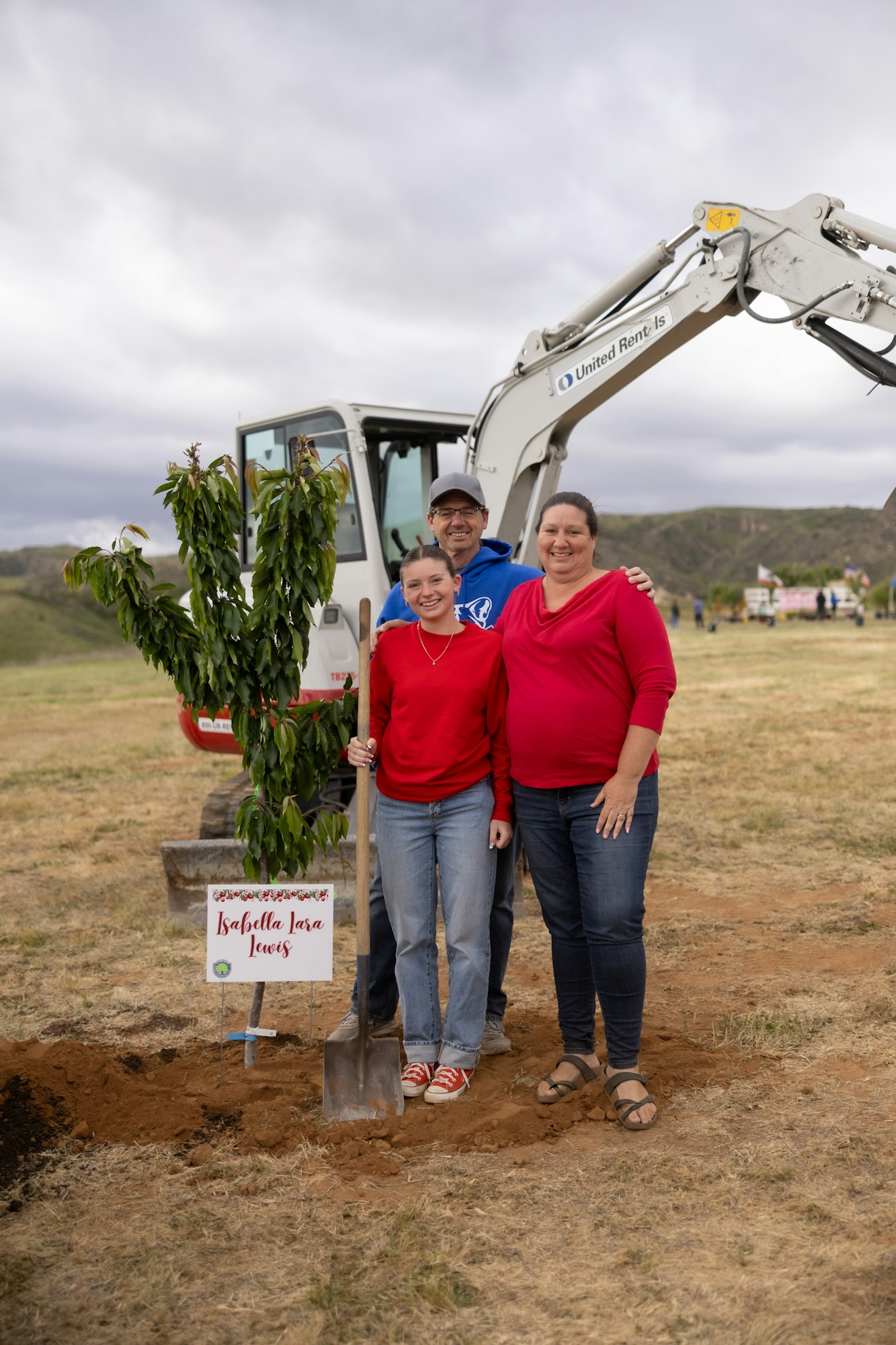 Three people with a tree ready to plant, a sign, and an excavator in the background on a cloudy day.