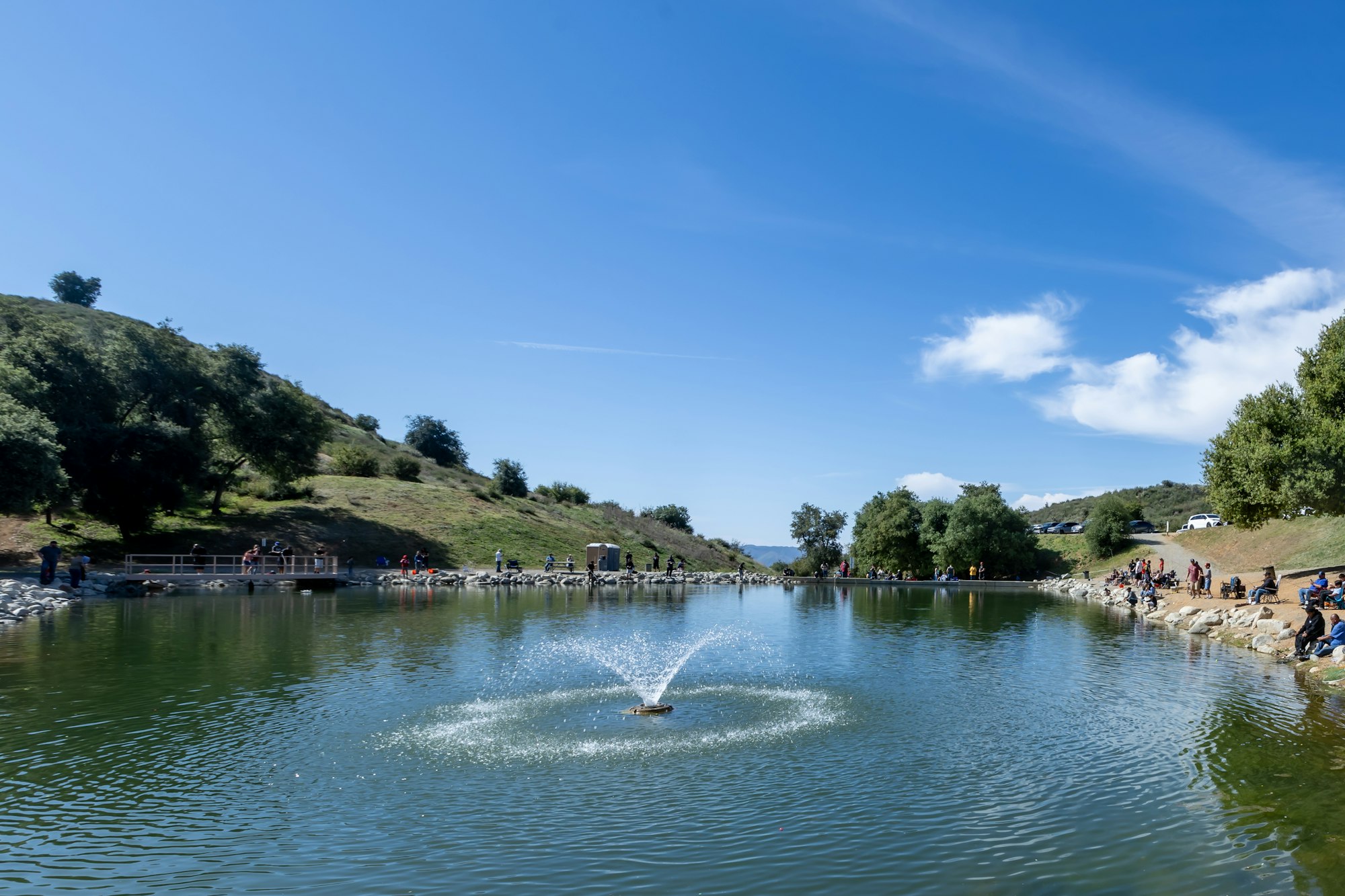 People relaxing by a pond with a fountain, surrounded by hills and trees under a blue sky.