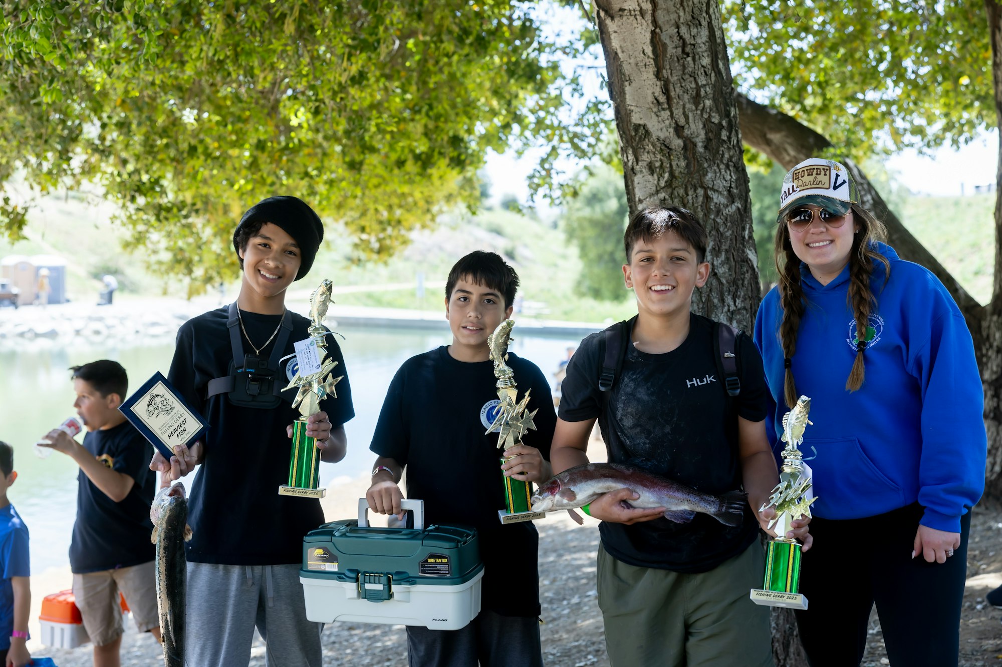 Four people smiling, holding trophies and fish, outdoors by a tree.