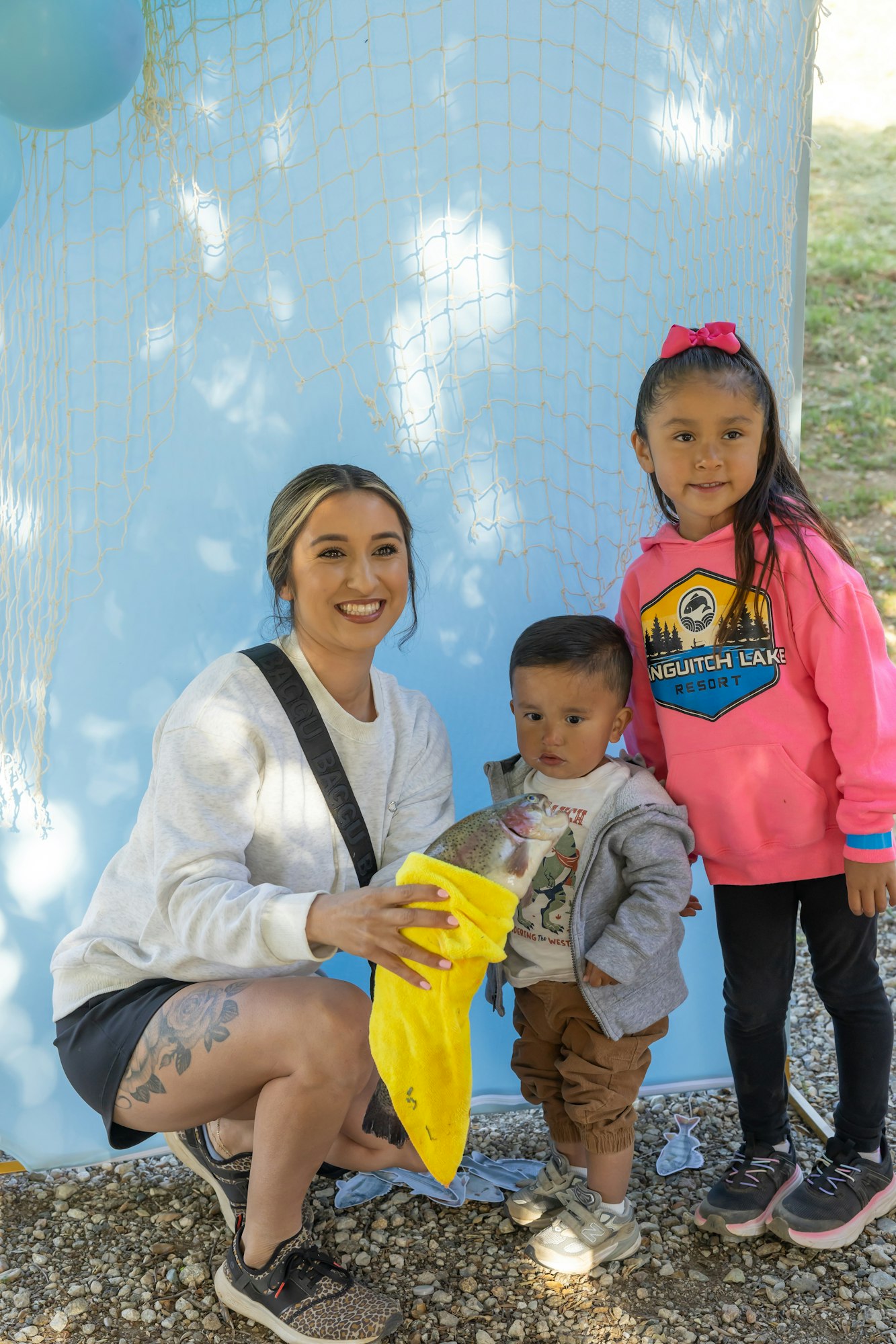 Three people smiling, holding a fish with a yellow cloth, in front of a blue backdrop with netting and balloons.