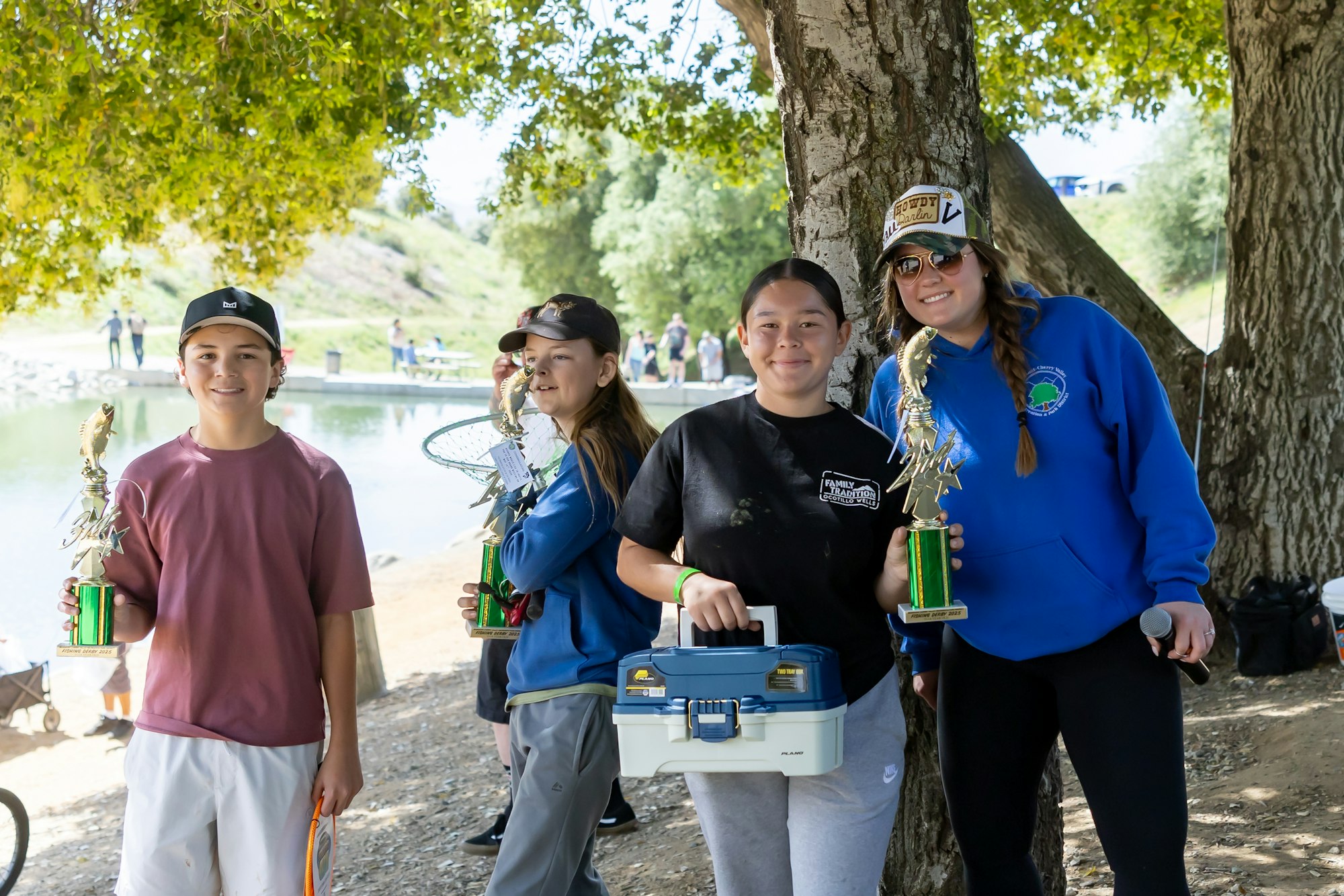 Four people holding trophies and a toolbox under a tree by a lake.
