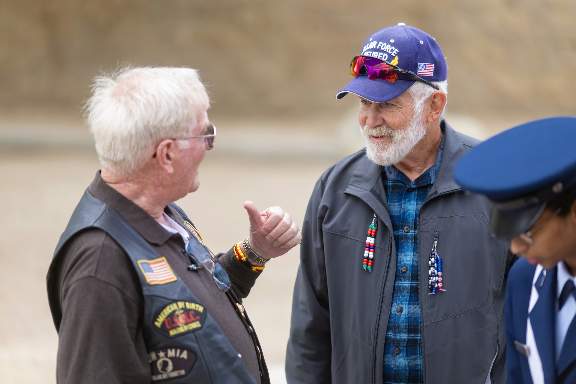 Three people, including two older men in jackets and caps, talking. One man's cap reads "Air Force Retired."