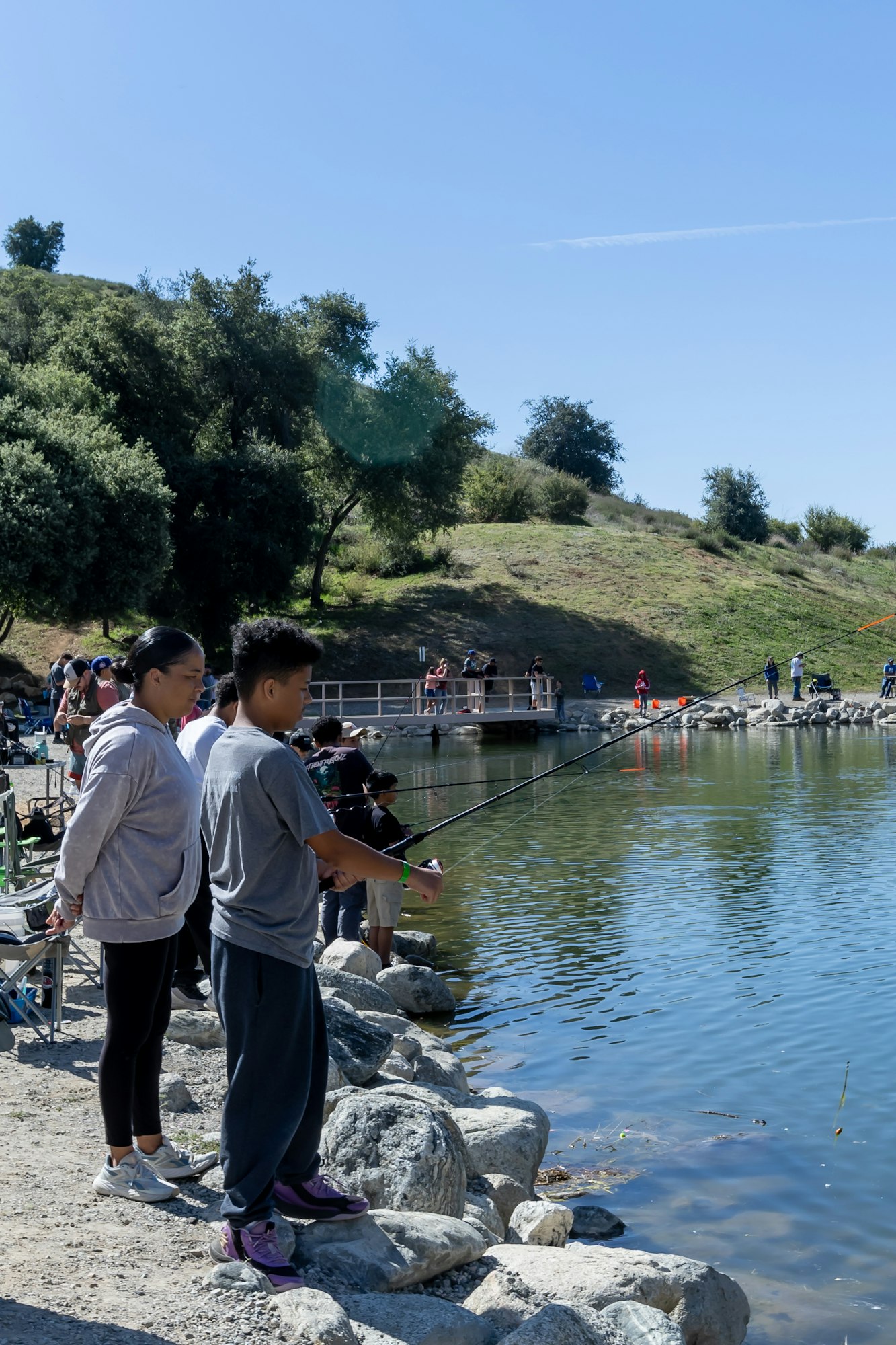 People fishing by a lake, surrounded by trees and hills under a clear blue sky.