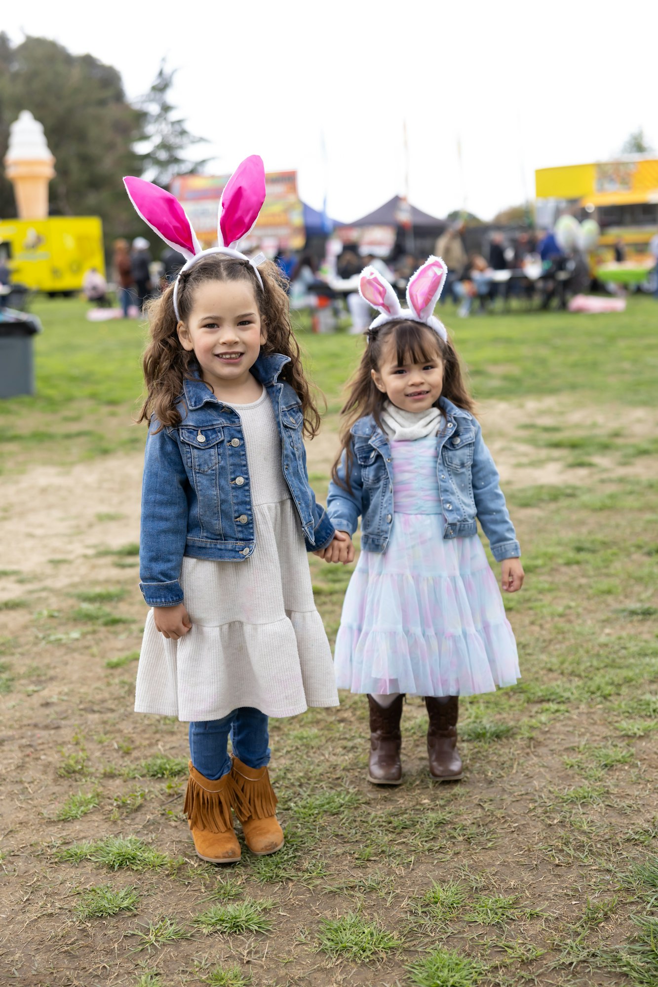 Two young girls wearing pink bunny ears and denim jackets, holding hands in a park.