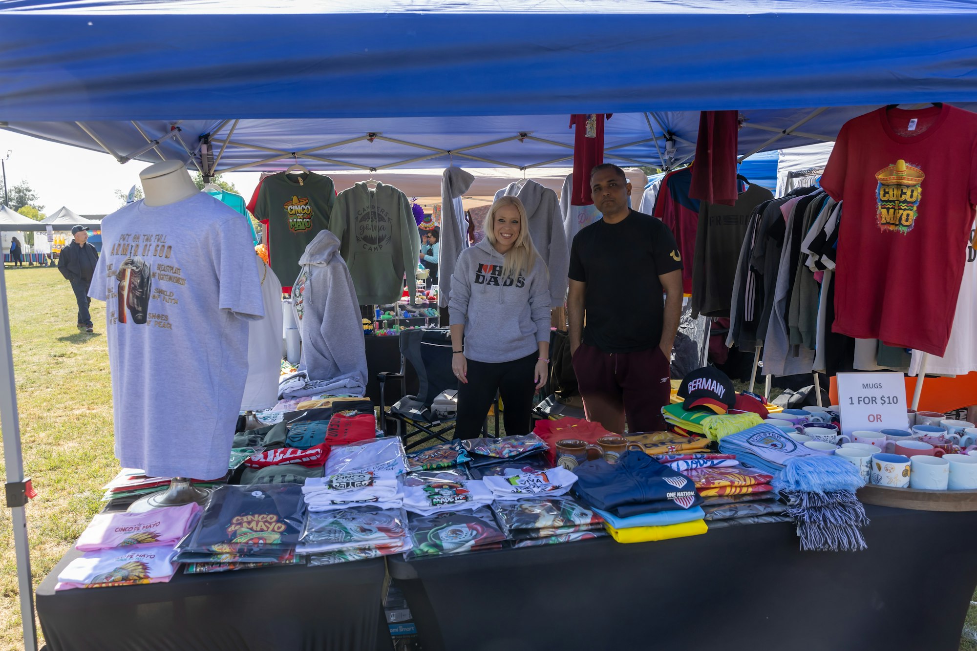 Outdoor market stall with T-shirts, sweatshirts, hats, and mugs. Two people smiling under a blue canopy.