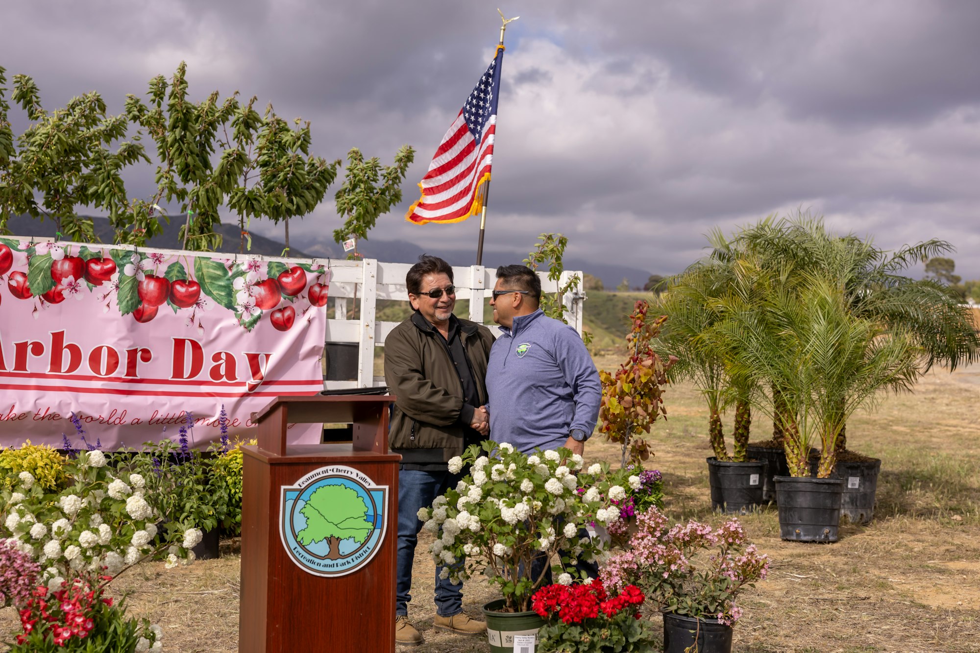 Two people shaking hands at an Arbor Day event with a banner, plants, and an American flag in the background.