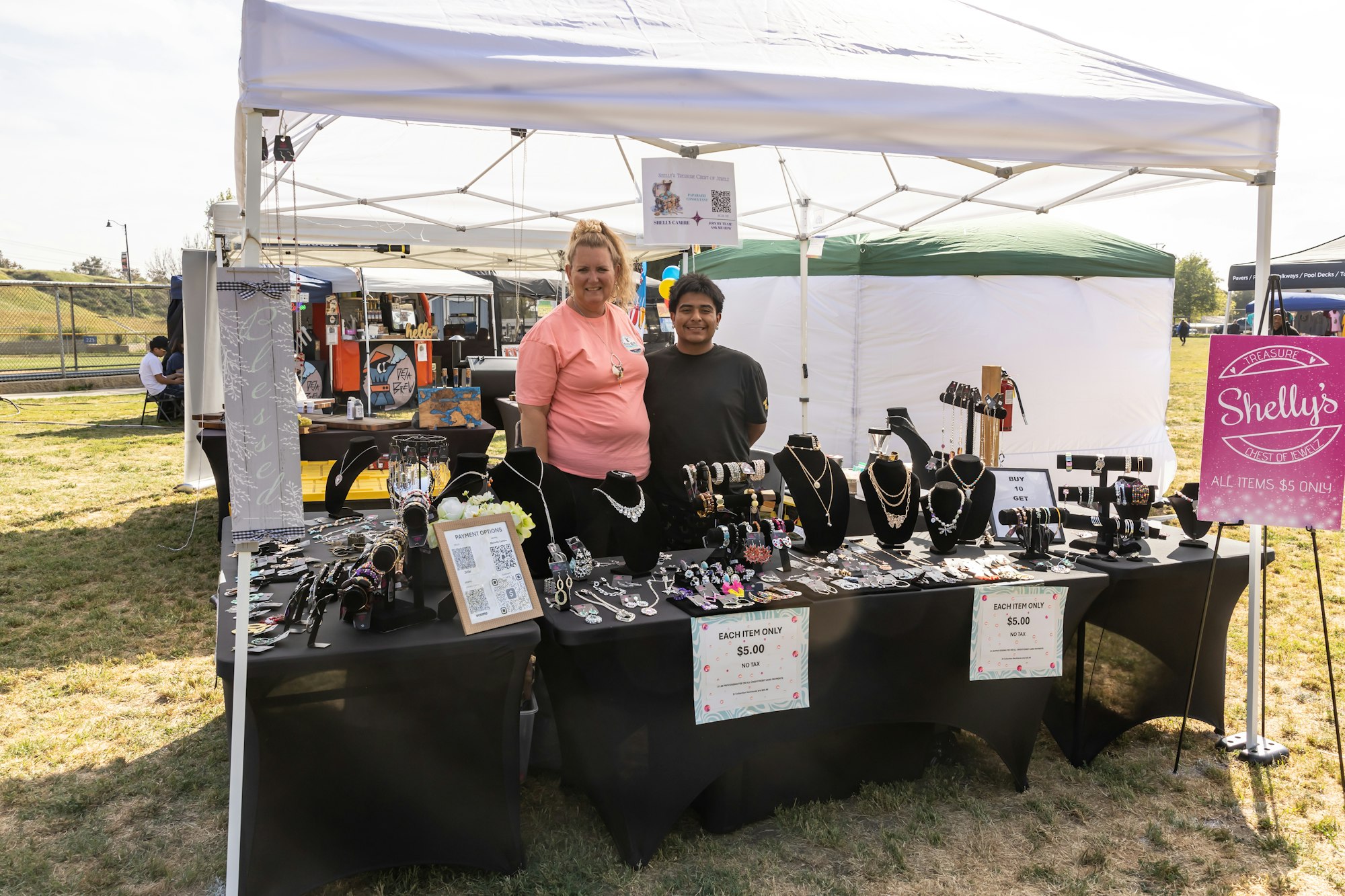 Two people stand at a jewelry stall with a Shelly's sign at an outdoor market.