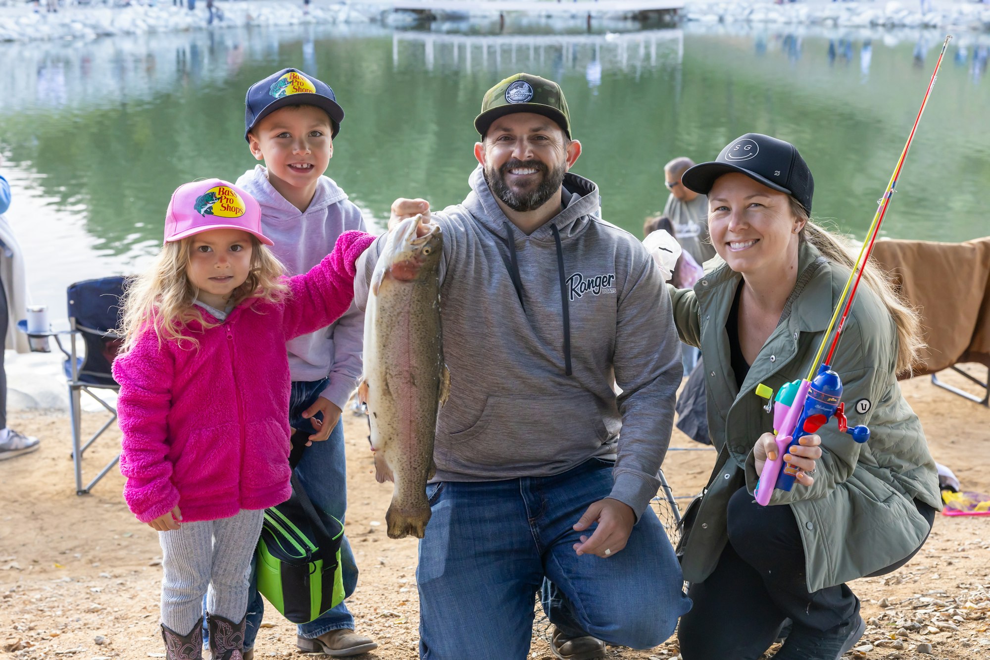 A family with two kids and two adults poses by a lake, holding a large fish and a colorful toy fishing pole.
